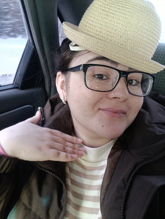 A woman with dark hair and glasses wears a straw hat perched atop her head. She is wearing a striped beige and white sweater and a dark brown jacket. Her left hand is raised near her neck. She has fair skin and small earrings. The woman is in a car, and the window is visible in the background.