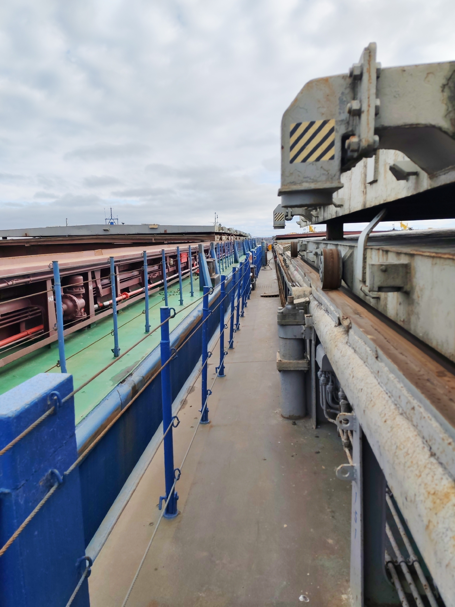 The image shows a walkway on a ship or industrial facility, with safety railings on the left painted blue, and industrial machinery on the right. The walkway is a light grey color. The machinery on the right has a warning sticker with black and yellow diagonal stripes. Additional industrial equipment is visible beyond the walkway. The sky is overcast. There is no verifiable evidence from this image to specify its location as Rybinsk, Russia.
