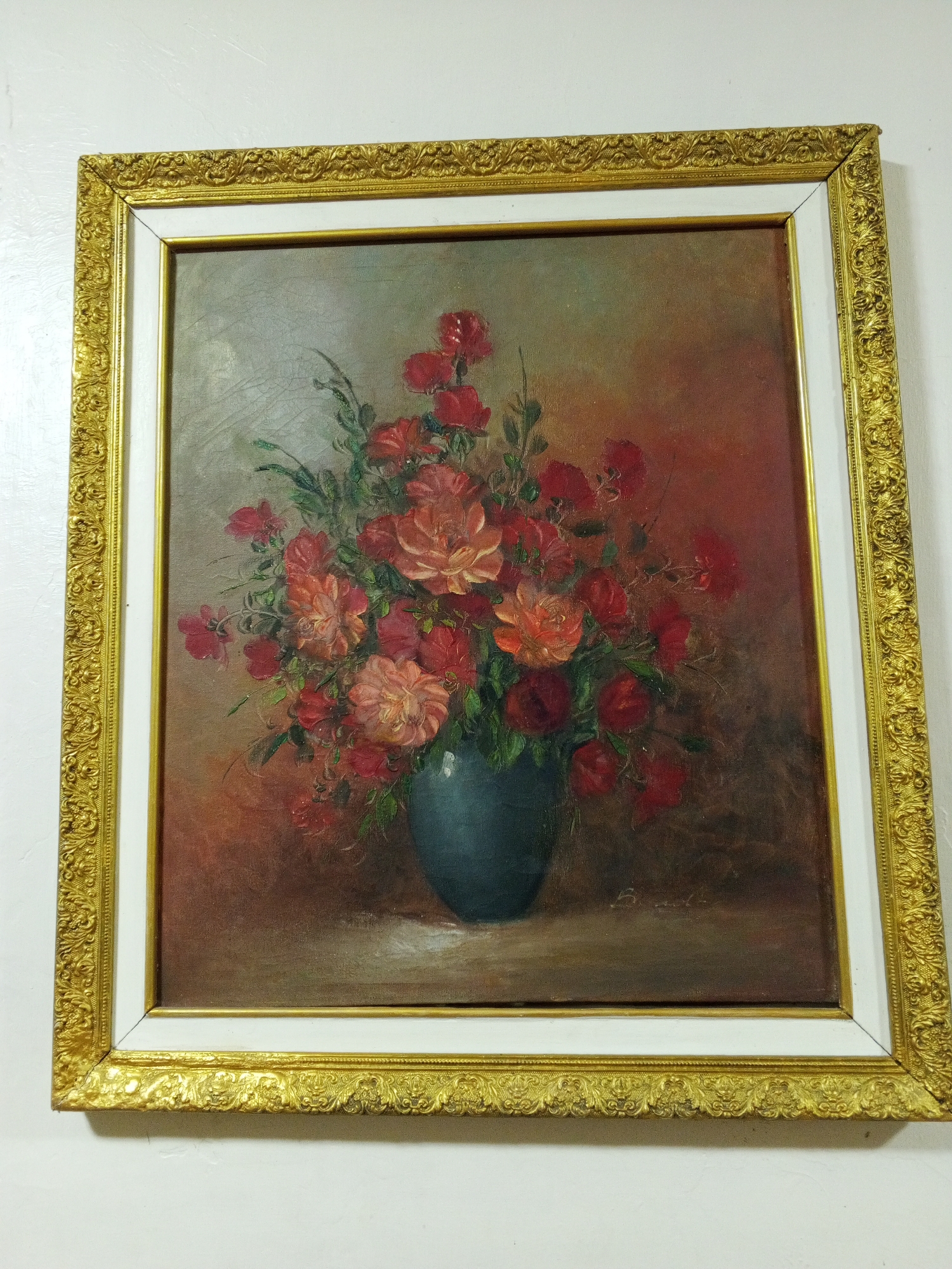 A framed painting hangs on a white wall. The painting depicts a floral arrangement in a blue vase. The flowers are primarily red and pink, set against a warm, reddish-brown background. The painting is framed with a gold, ornate outer frame and a white inner frame. The location of the image is the city of Tinaquillo, Venezuela. There are no people in the image.