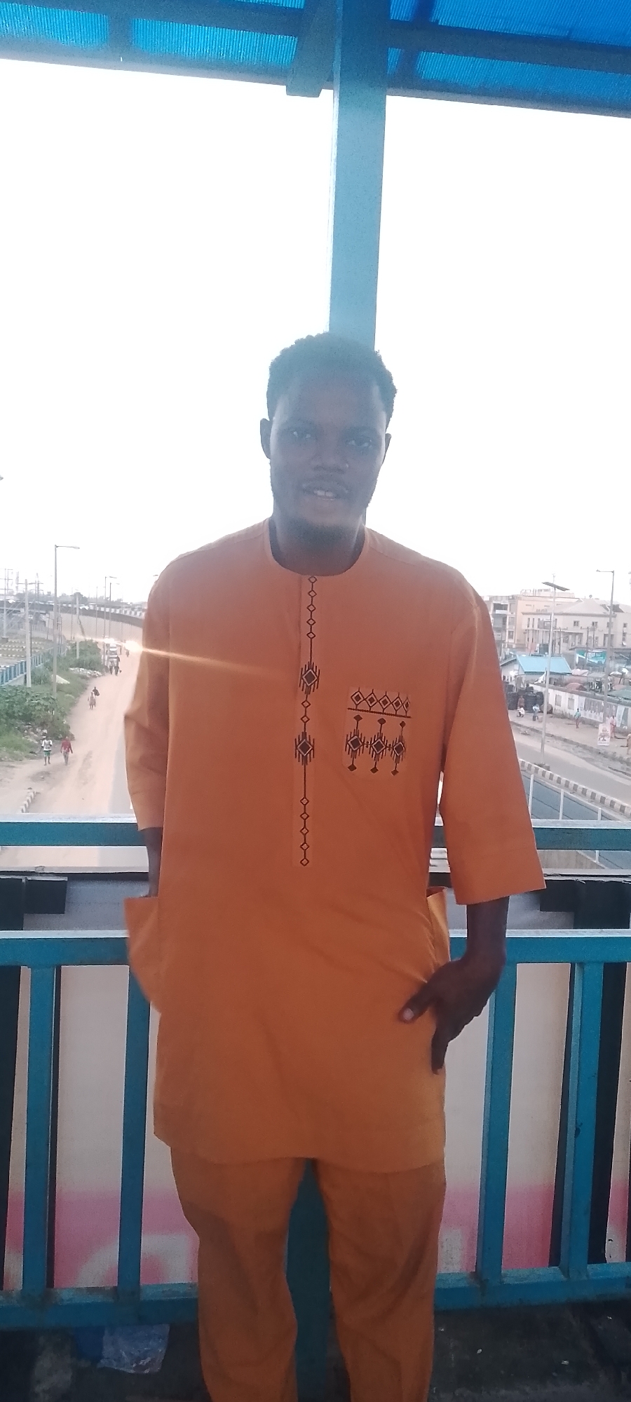 A young Black man stands on a pedestrian bridge, looking towards the camera with a slight smile. He is wearing a bright orange traditional African outfit with intricate black embroidery on the chest. The bridge has a blue railing and a blue-tiled roof overhead. In the background, a busy street with cars and pedestrians can be seen, along with buildings and power lines, suggesting an urban environment. The sky is bright and hazy, indicating daylight.