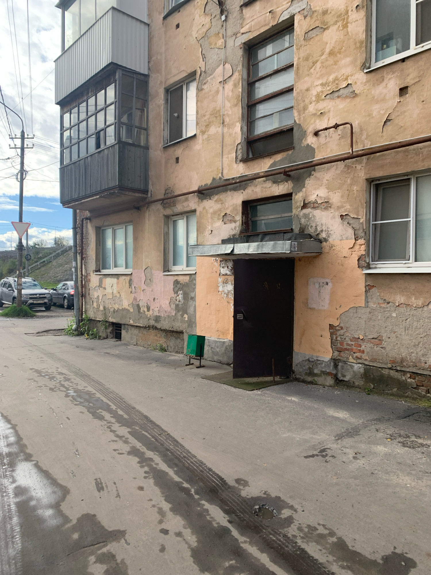 The image depicts the exterior of a weathered, multi-story building in Arkhangelsk, Russia. The facade shows significant peeling paint and exposed brickwork, with a balcony on the upper floors featuring a dark wooden enclosure with multiple glass panes. Below the balcony, a dark metal door is visible, set within an entrance alcove. A green trash bin stands to the right of the entrance. The building is situated adjacent to a paved road with tire tracks and wet patches, suggesting recent rain. In the background, two cars are parked on the side of the road, and a traffic sign can be seen on a pole. The sky is partly cloudy. The overall scene conveys a sense of urban decay and the realities of life in a northern Russian city.