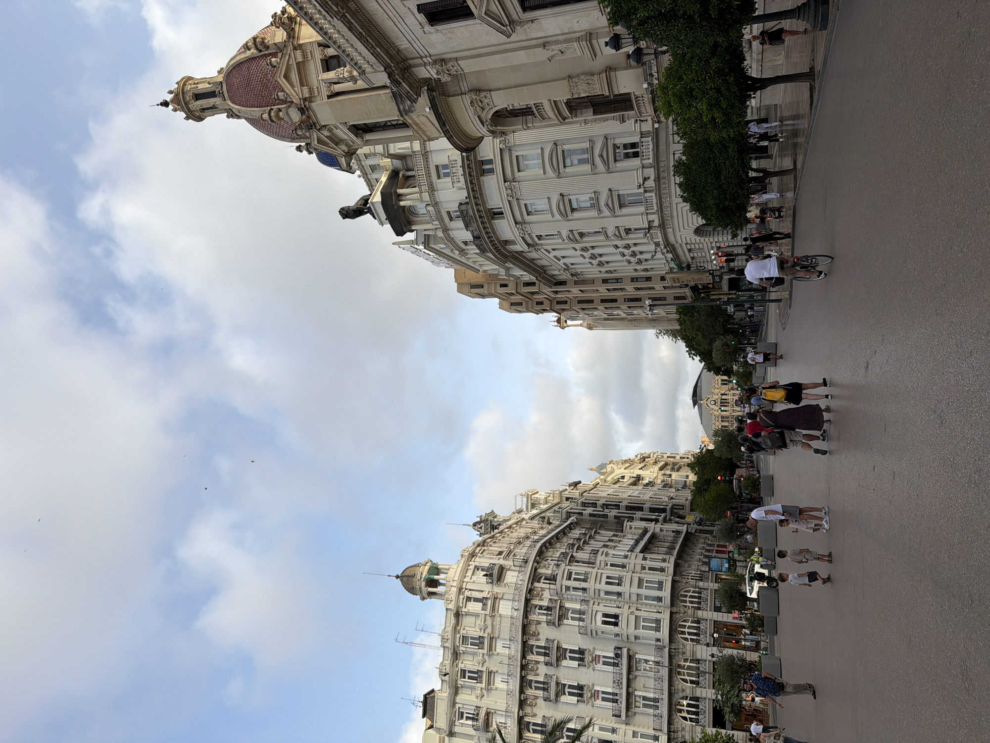 In this image, a wide, paved plaza is captured in an urban environment. On the left and right sides of the plaza, grand, multi-story buildings with intricate architectural details line the street. The buildings feature numerous windows, balconies, and decorative elements, with one building on the right topped by a distinctive red-domed cupola. The sky above is a mix of light blue with scattered clouds, and several birds are visible in the upper portion of the frame.
In the plaza, various people are present. On the left, a man in a blue shirt walks away from the camera, while a group of people, including children, are gathered in the center. Further into the plaza, a cyclist rides on the road. Other individuals are seen walking on the sidewalks and in the distance, suggesting a public space with moderate pedestrian activity. Trees are planted along the edge of the buildings, providing some greenery. The overall impression is a European city square with significant historical architecture.