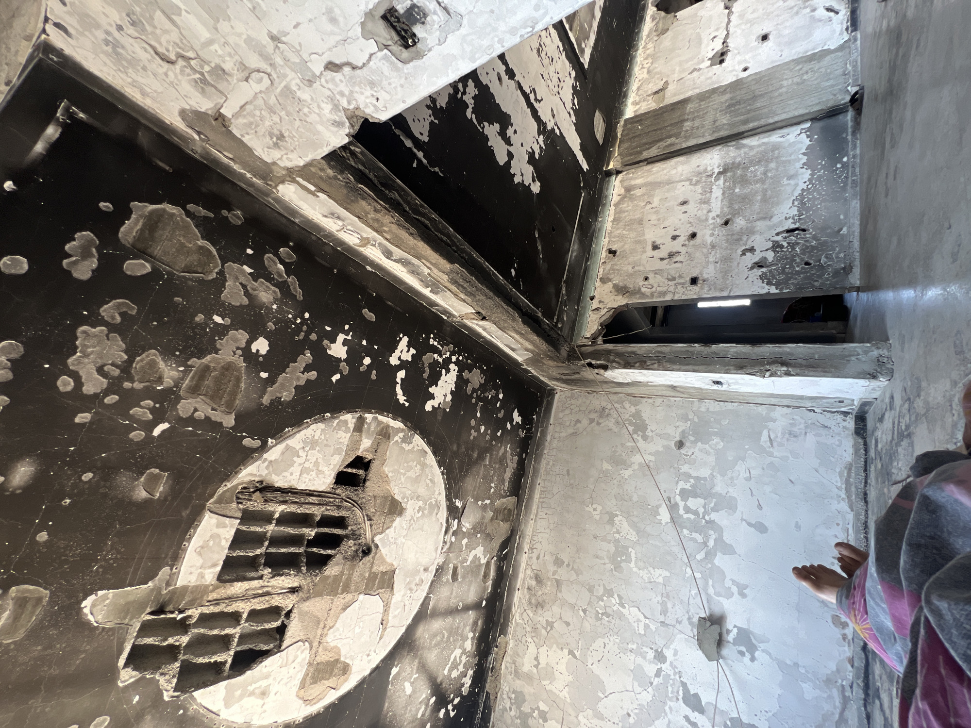 This image depicts the interior of a severely damaged building in Gaza. The ceiling is heavily charred and peeling, with a large circular section in the upper left corner showing exposed concrete with a grid-like structure. The walls are rough and discolored, with patches of plaster missing and evidence of impacts, possibly from shelling, visible as numerous small holes. The floor appears to be concrete and is uneven. In the foreground, a person is lying down, covered by a patterned blanket, with their feet visible. They are covered by a blanket in shades of pink, grey, and yellow. The overall impression is one of destruction and neglect within an urban environment.
