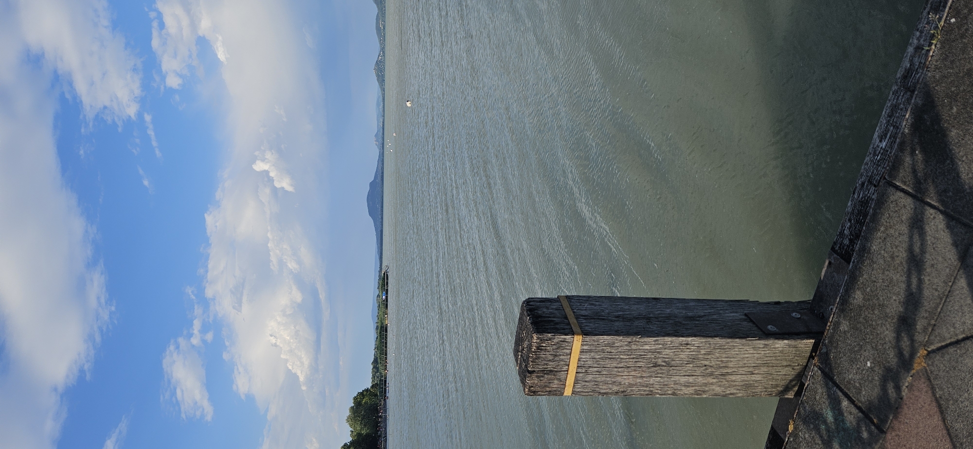 The image depicts a calm lake scene with a wooden bollard in the foreground. The bollard, made of weathered grey wood, has a yellow band around its top and is bolted to a stone pathway. The pathway, constructed of dark grey and reddish paving stones, leads to the edge of the lake. The lake water is a muted green, with gentle ripples creating a textured surface. In the mid-ground, a thin wooden pier or walkway extends into the water, lined with reeds and vegetation on the left bank. Beyond the pier, distant land formations are visible, including trees on a far shore and a prominent, cone-shaped mountain in the distance. The sky above is a vibrant blue, dotted with fluffy white clouds. The overall scene suggests a peaceful daytime environment. This location is identifiable as the Balaton shore in Balatongyörök, Hungary, a popular tourist destination known for its scenic views of Lake Balaton and the surrounding landscape, including the Tapolca Basin.