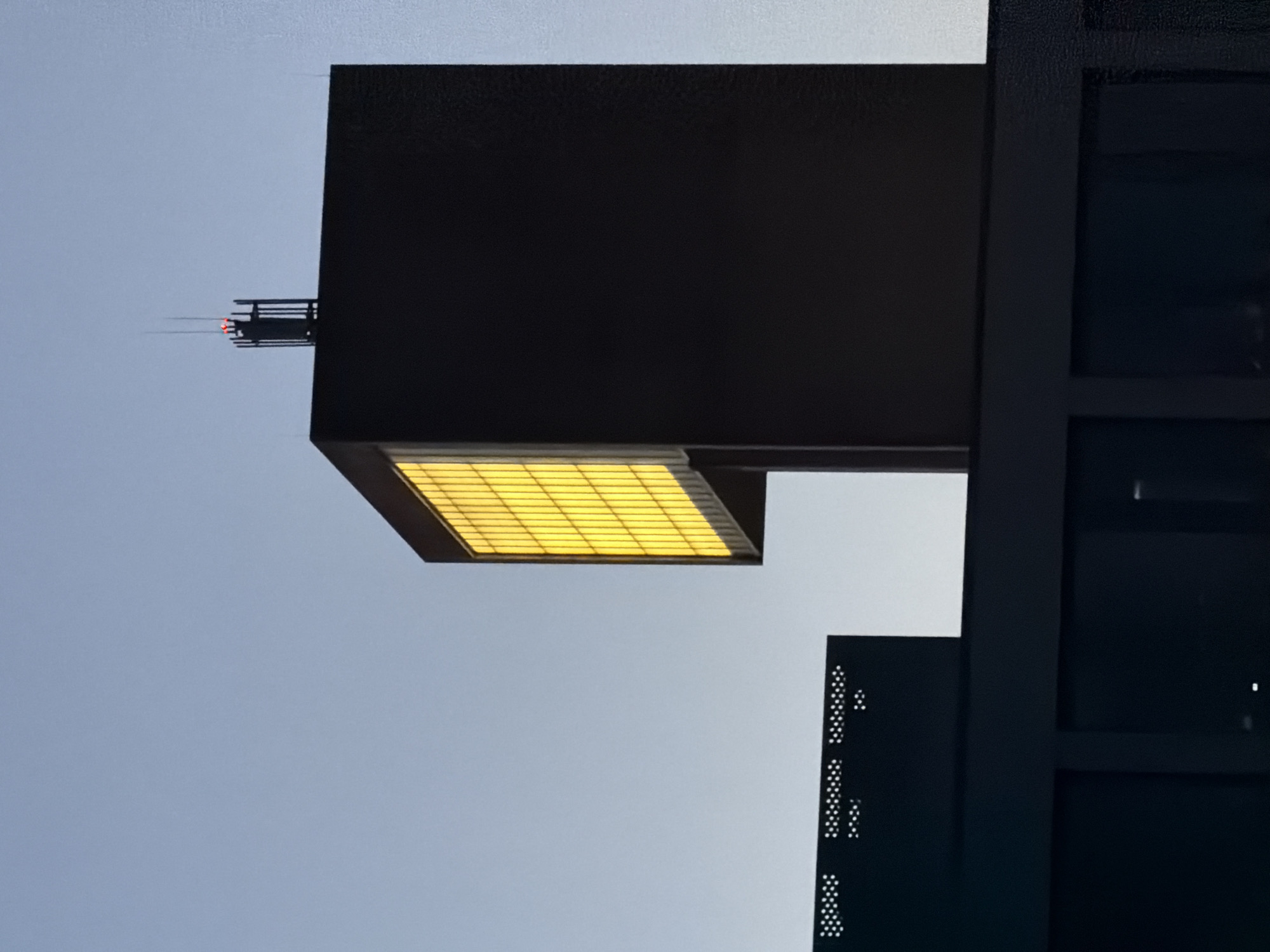 The photograph captures a modern architectural structure in Leudelange, Luxembourg, against a clear, pale blue sky. The dominant element is a dark, rectangular building with a prominent, illuminated yellow facade composed of grid-like panels. This building is topped with a dark structure that features two thin vertical antennas, one of which has a small red light at its apex. In the foreground, a lower, dark structure with a perforated pattern is visible, suggesting a larger complex. The overall scene is minimalist and stark, emphasizing the geometric shapes and the contrast between the dark materials and the luminous yellow window. No people are visible in the image. The lighting suggests it is either dawn or dusk.