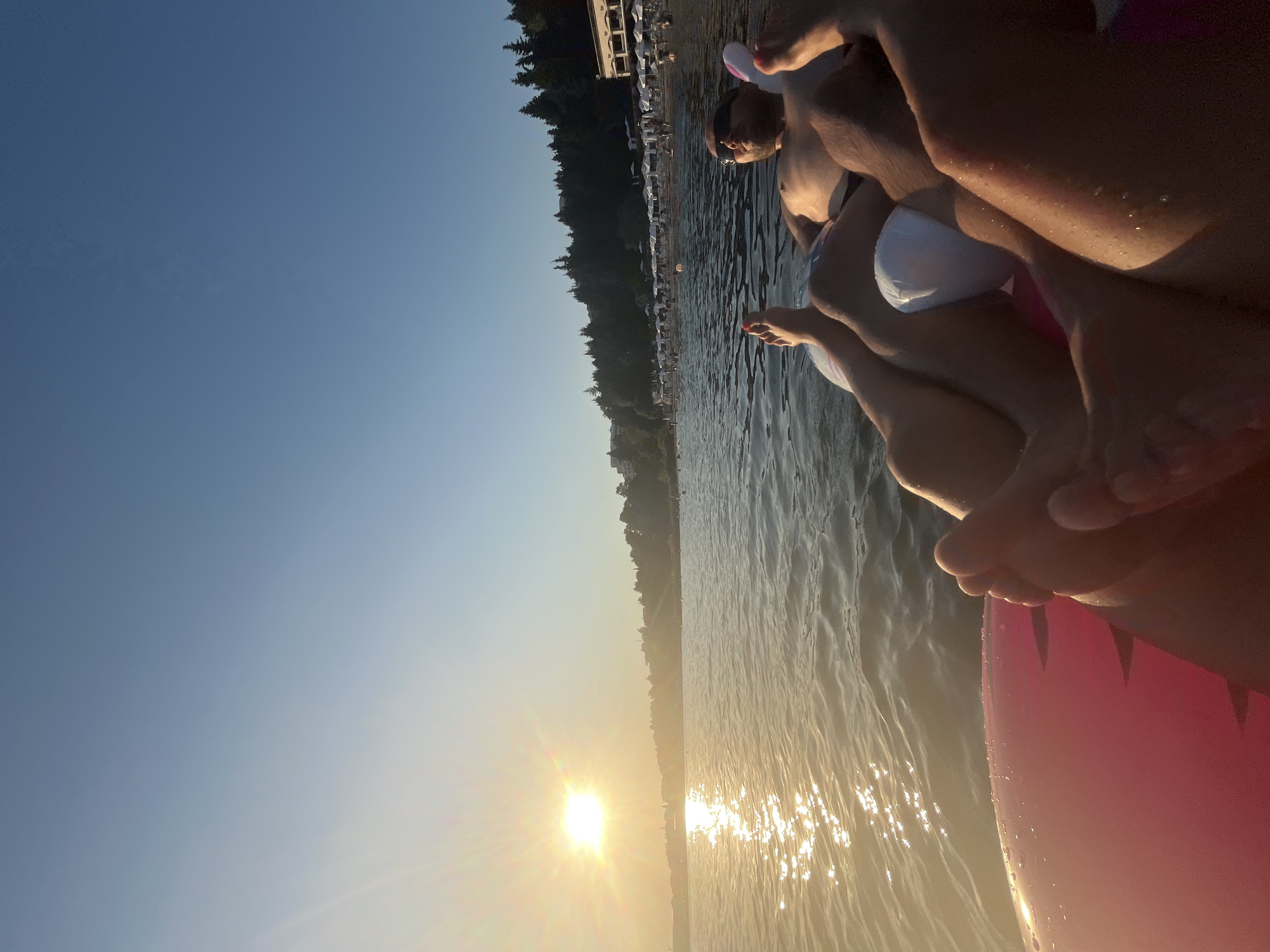 Two individuals are relaxing on an inflatable raft in the calm waters of the Black Sea, facing a beach lined with white umbrellas and lush green trees. The sun is setting on the left side of the image, casting a warm, golden glow on the water and the sky, which transitions from a pale blue near the horizon to a deeper blue above. The shoreline in Nesebar, Bulgaria, features a few buildings nestled among the trees and a stretch of sand dotted with beachgoers. The perspective is from the water, looking towards the shore, with the raft and the legs of the two people prominently featured in the foreground.