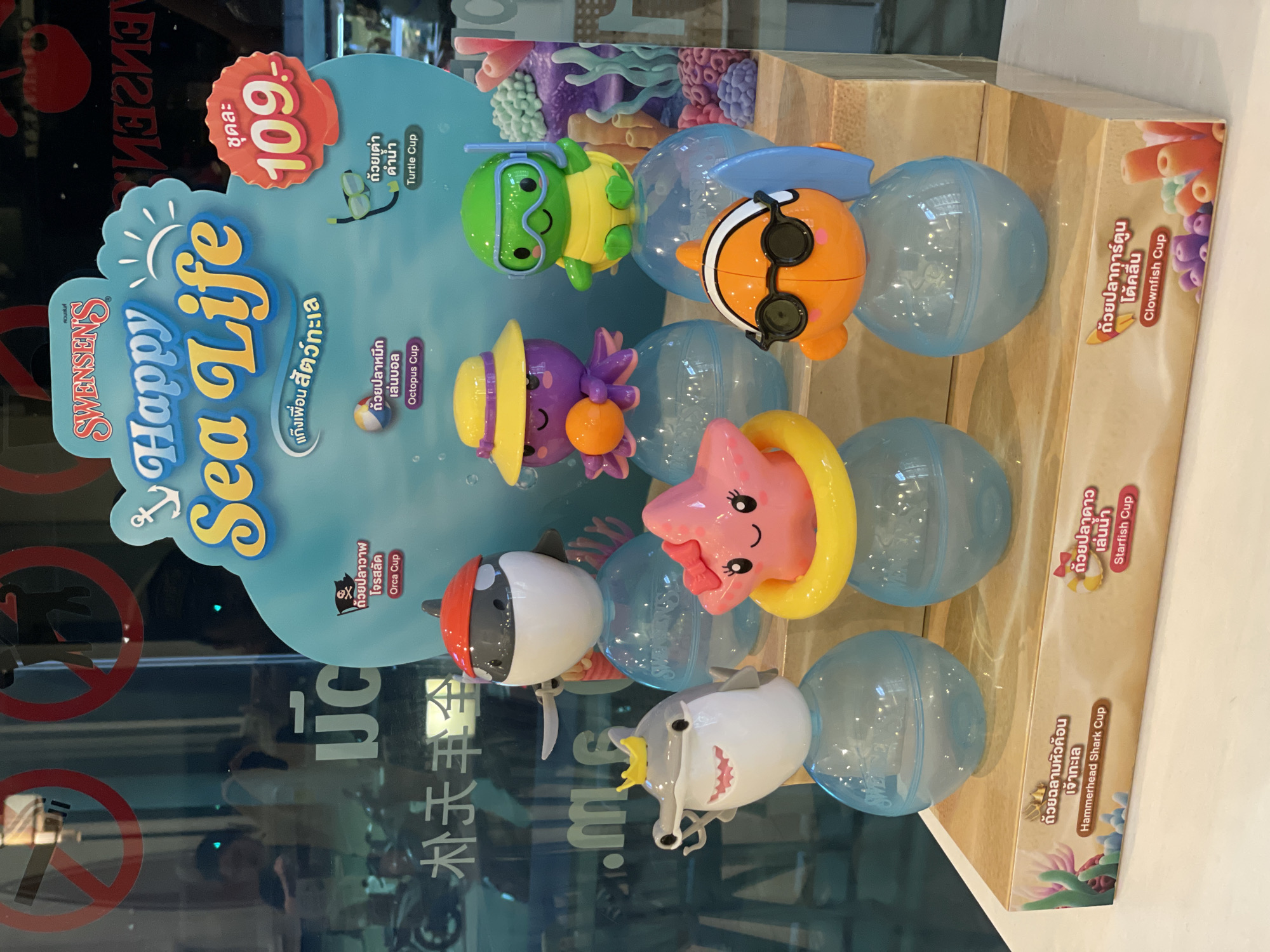 This image shows a display of Swensen's Happy Sea Life collectible cups in what appears to be a store or restaurant setting. The display features a stand with a light blue background and various sea creature cup designs: Turtle, Clownfish, Octopus, Starfish, Orca, and Hammerhead Shark. Each cup is displayed on a light blue plastic ball. The price tag on the display reads "109.-" in Thai baht per cup. The display is located inside a building, as seen through the glass reflection, with signage in Thai and Chinese visible. Given the brand Swensen's and signage details, the location is likely in Pattaya City, Thailand.