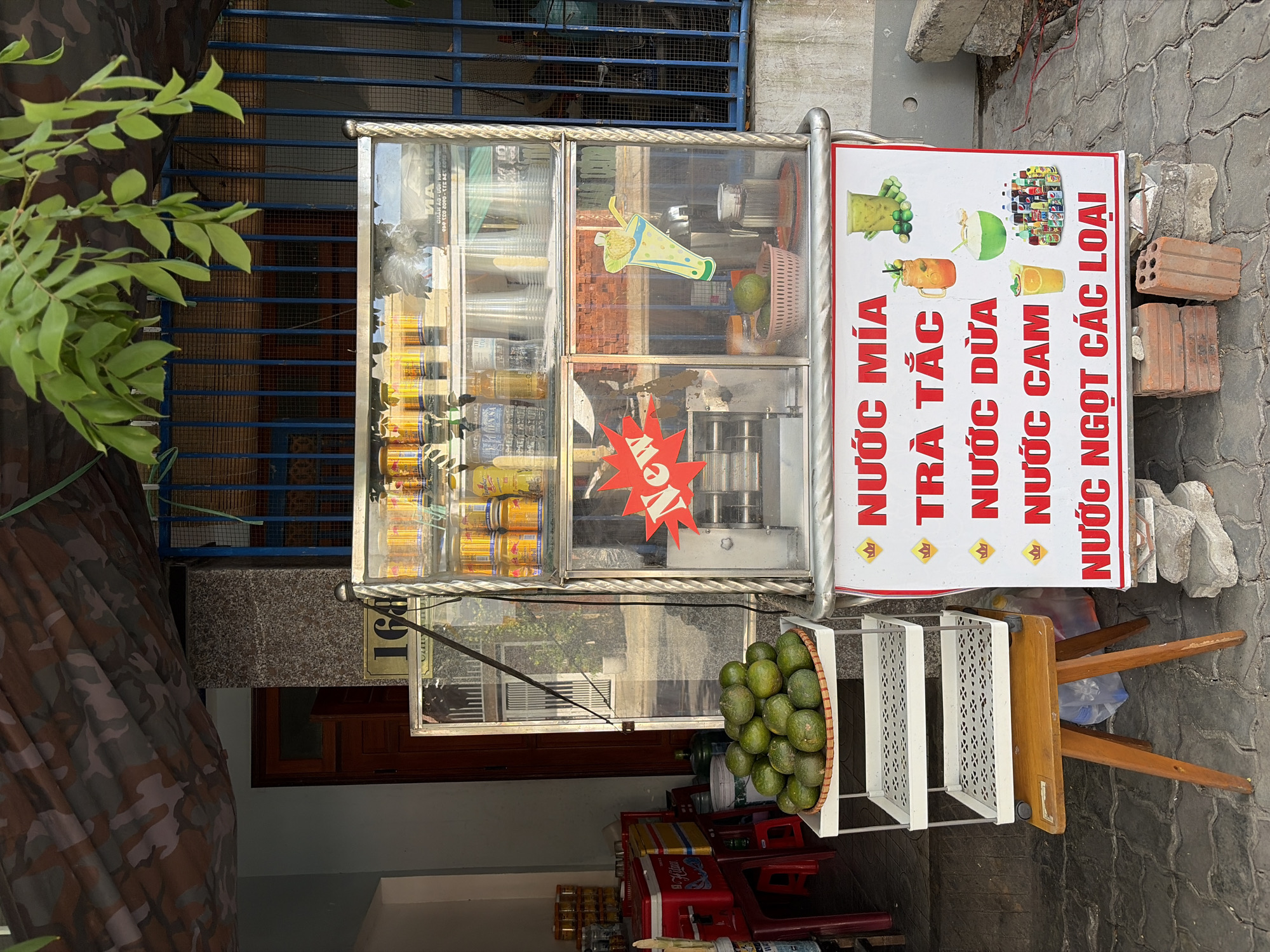 This is a street-level, eye-level medium shot of a juice stand in Da Nang, Vietnam. The stand is made of glass and metal and displays various canned drinks, cups, and fruit, including a basket of green limes. A sign on the front of the stand lists different juices like "Nước mía" (sugarcane juice), "Trà tắc" (kumquat tea), "Nước dừa" (coconut water), and "Nước cam" (orange juice). There are no people visible in the image. The stand is positioned on a paved sidewalk in front of a building with a camouflaged awning. The environment appears to be an urban street scene.
