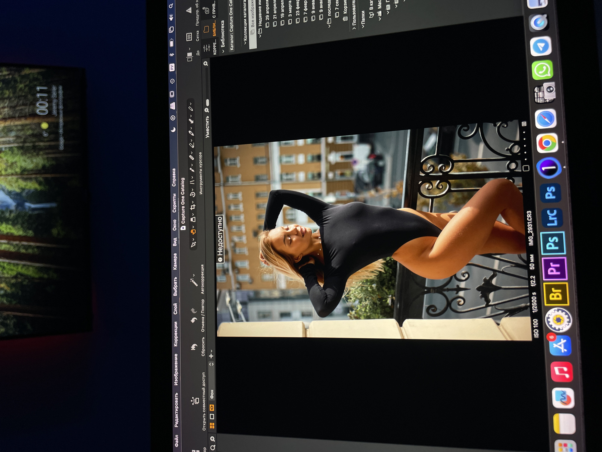 The image is a photograph of a laptop screen displaying the Capture One photo editing software with a woman in a black bodysuit posing on a balcony. The woman is blonde and is lying down. Her body is turned to the side, facing the viewer. The background shows an urban environment, presumably a building with windows and architecture characteristic of city. The laptop's interface shows menus and editing tools related to the software, and the dock at the bottom contains various icons. The photograph is taken indoors, possibly at night or in a dimly lit room, as indicated by the laptop screen's brightness compared to the surroundings. The display clock and date can be seen on the left side of the screen. The location in the city of Kazan, Russia is suggested by the Cyrillic script used for software menus and interface elements.