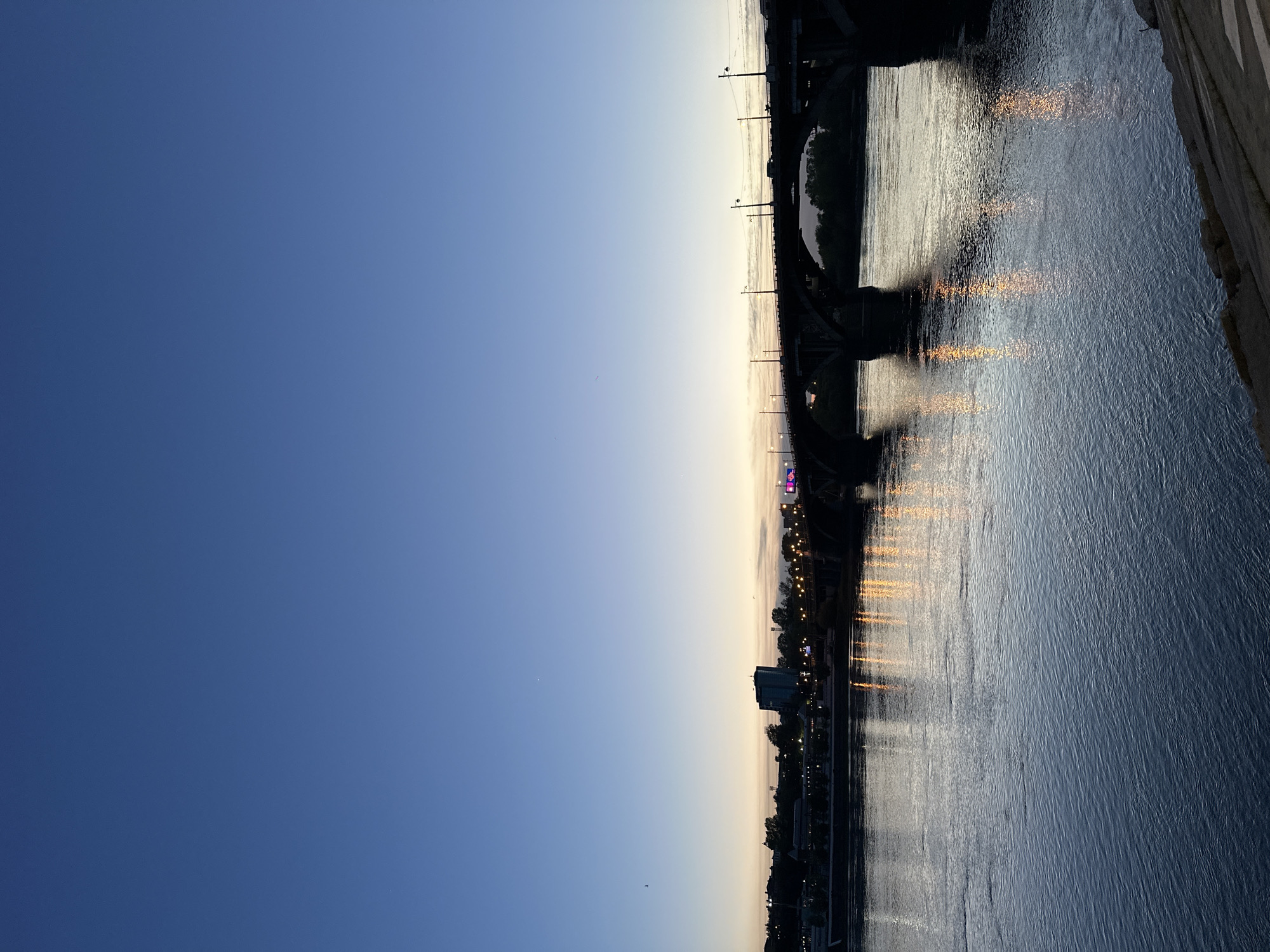 The image depicts a horizontal view of the Angara River in Irkutsk, Russia, at dusk or dawn. The sky transitions from dark blue to a lighter, faded yellow-orange toward the horizon. The river reflects the city lights, creating vertical streaks of warm light on the water's surface. Silhouettes of buildings and trees line the riverbank. A bridge spans the river, with its structure partially visible against the fading sky. The river's surface shows some ripples and texture.