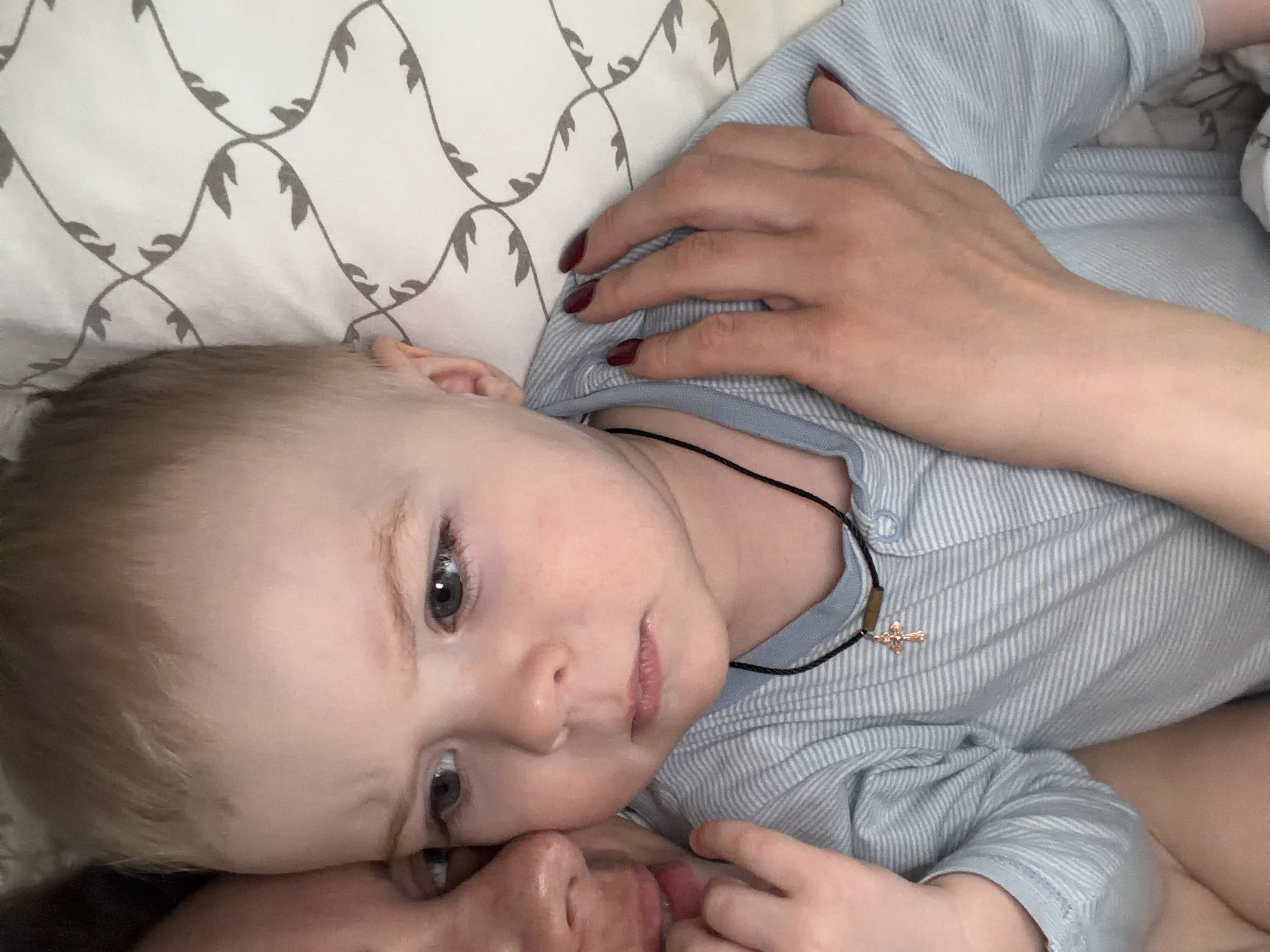 A baby lies on top of an adult's chest in bed. The baby is fair-skinned with blonde hair and grey eyes. They wear a light blue striped shirt and a black cord necklace with a copper-colored cross pendant. The adult is positioned beneath the baby with their face partially visible near the bottom of the frame. One of the adult's hands rests on the baby's chest. The background consists of a white pillow with a repeating grey leaf pattern. There is no information in the image to indicate that the location is Votkinsk, Russia.