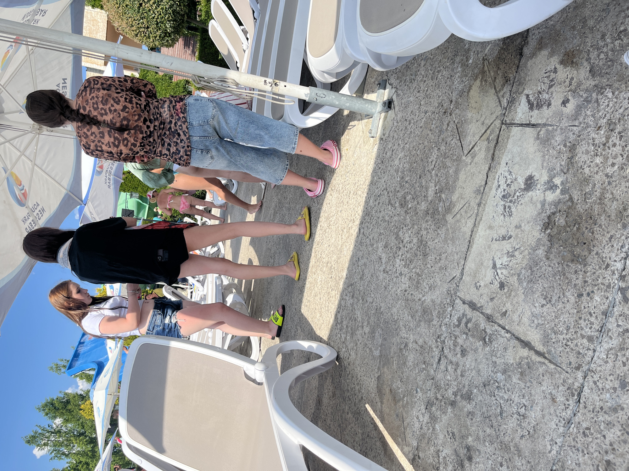 Three young women stand near lounge chairs in an outdoor area, possibly a water park or resort, under a large umbrella. The woman on the left has long brown hair and wears a white t-shirt and denim shorts. The woman in the middle has dark hair and wears a black top and black shorts. The woman on the right has dark hair in a braid and wears a leopard-print top and denim capris. She also wears pink Crocs. The ground is paved with concrete slabs. In the background, there are lounge chairs, trees, and a blue waterslide. The sun is shining brightly. The scene is likely located in Ravda, Bulgaria, a popular resort town on the Black Sea coast known for its beaches and water parks.