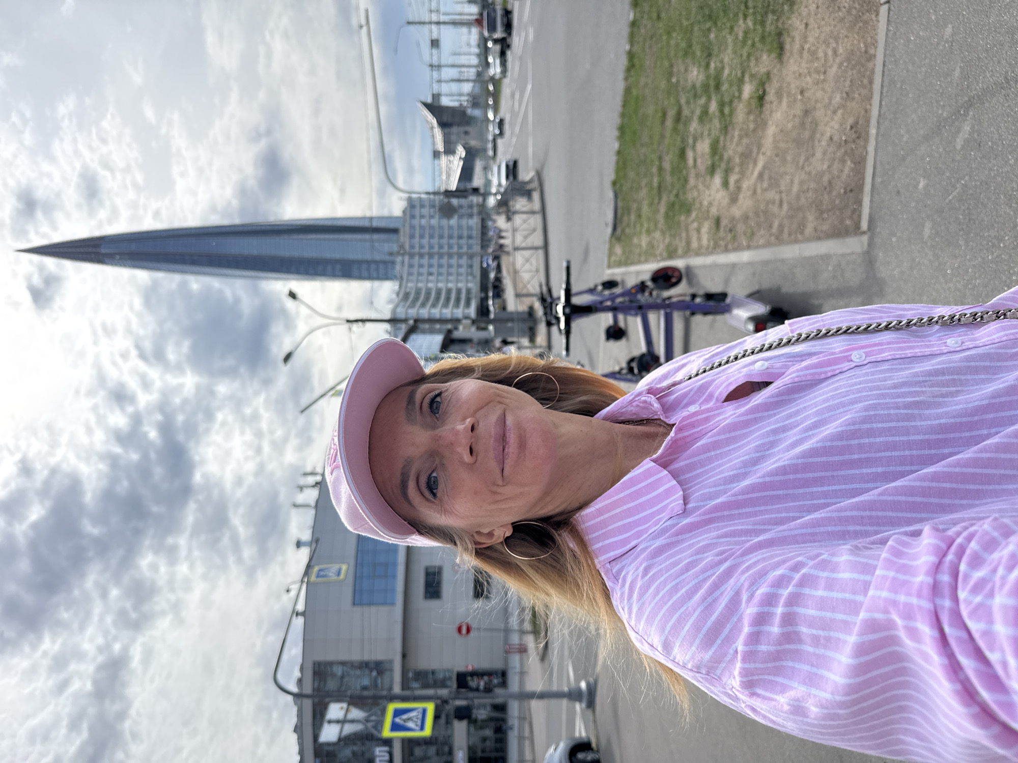 A medium eye-level shot depicts a fair-skinned woman with light brown hair wearing a pink-striped shirt and pink baseball cap, and gold hoop earrings. The woman is smiling toward the camera. In the background is an urban landscape with a tall, sharp-pointed building resembling a needle, presumably the Lakhta Center, a famous landmark in Saint Petersburg, Russia. Additionally, there are scooters, other buildings, a pedestrian crossing sign, street lamps, and vehicles visible in the scene. The sky is cloudy. The woman's proximity and focus imply that she took a selfie.