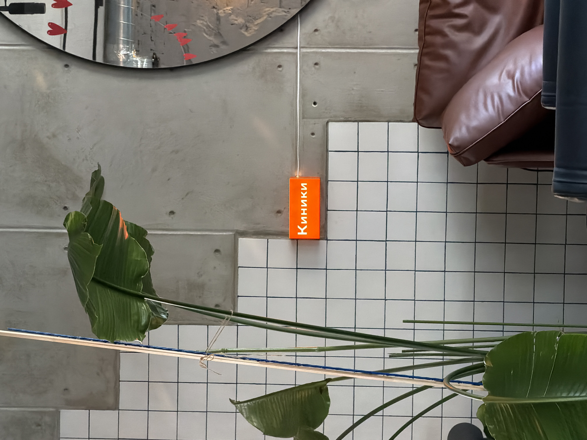 An orange illuminated sign reading "Киники" is mounted on a concrete wall. A large green leafed plant with a wooden support pole stands to the left of the sign. To the right of the sign, a round mirror with red heart and flag decorations is visible. A brown leather sofa is positioned in the lower right corner of the image. The wall features a grid pattern of white tiles. The overall scene appears to be an indoor setting, possibly a cafe or lounge.