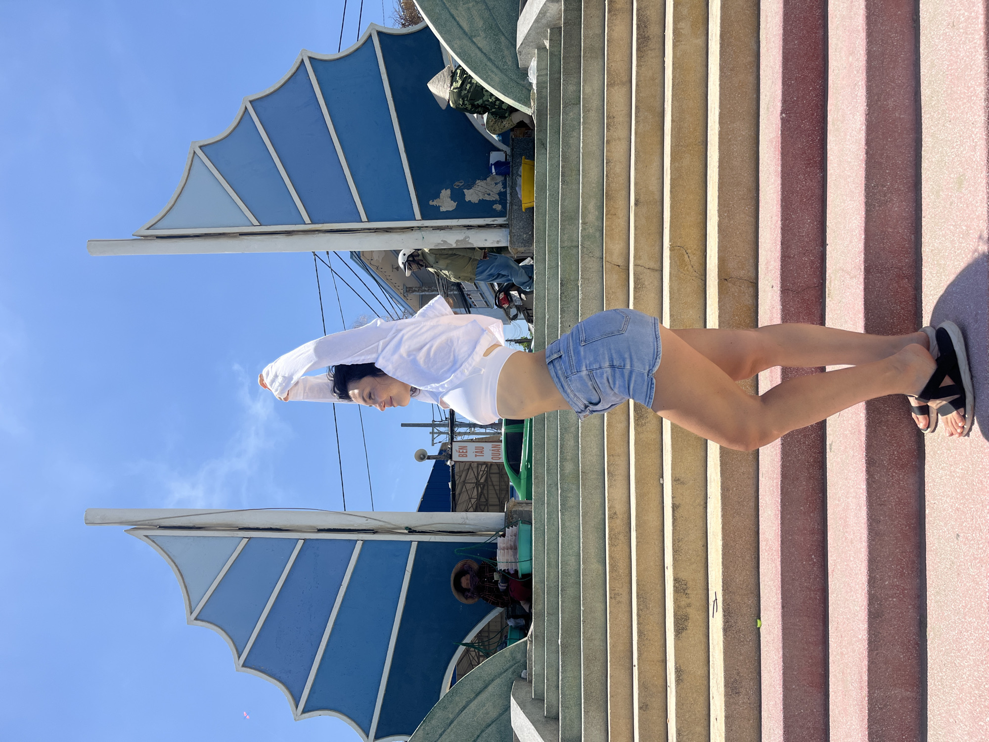 In the city of Mũi Né, Vietnam, a woman with dark hair, wearing a white crop top, denim shorts, and black sandals, stretches her arms above her head while facing right. She stands on a set of wide, outdoor concrete stairs, with some steps colored pink and others a sandy yellow. Behind her, a large, blue, sail-like structure with white accents rises against a clear blue sky dotted with faint clouds. To the left, a person in a conical hat sits on the stairs, attending to a collection of items in green baskets. Further back, a person in a green jacket and blue pants stands near a motorcycle, and another person in camouflage clothing and a conical hat is seated to the right. A green car is visible in the background, along with buildings and utility lines. The scene appears to be an outdoor public area, possibly a market or scenic overlook.