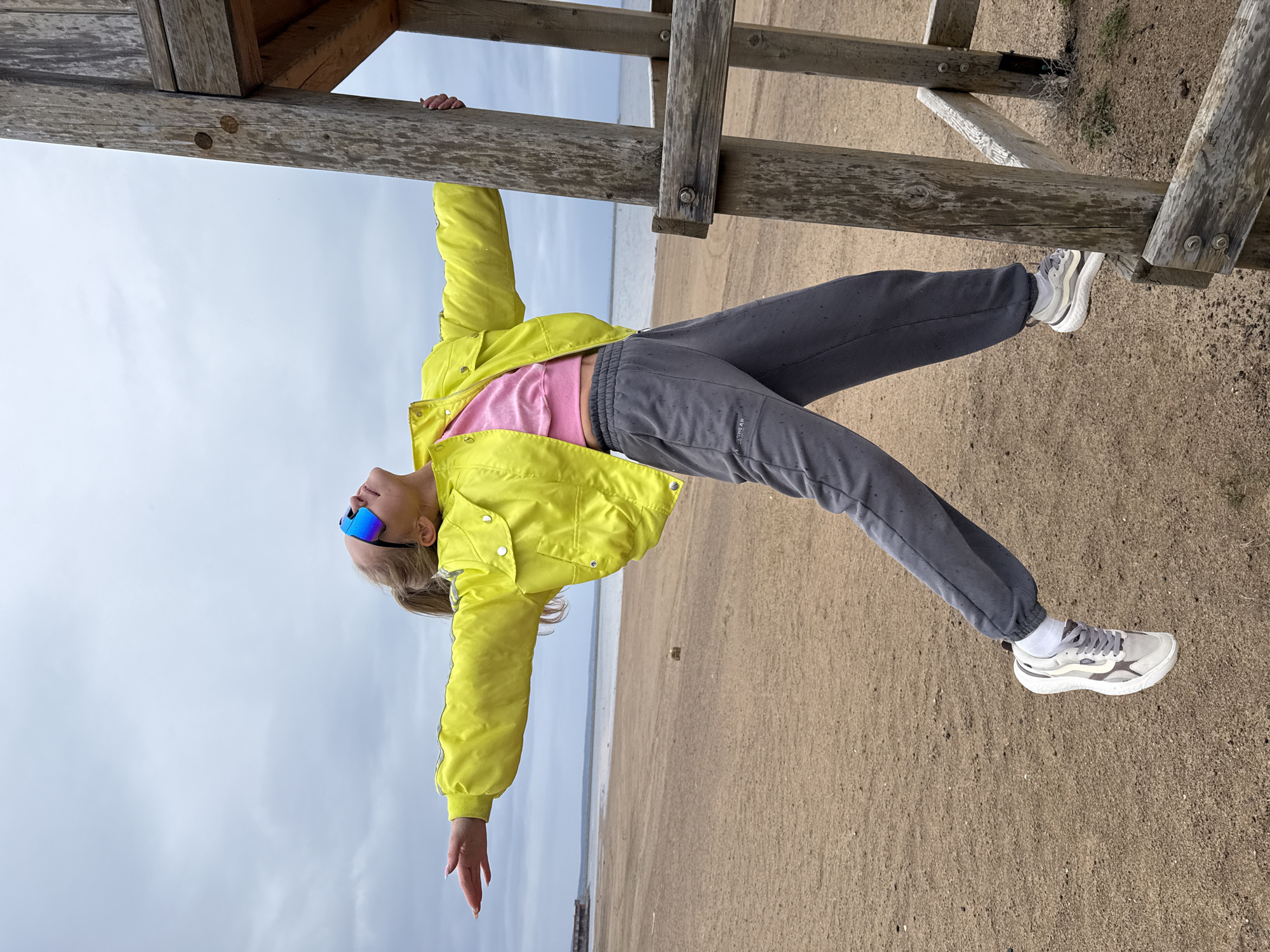 The original image is rotated 90 degrees counter-clockwise. It features a young woman suspended horizontally from a weathered wooden structure on a sandy beach. She is holding onto a thick wooden beam with both hands, with her body arched and her head tilted back, looking upwards. She has blonde hair pulled back and wears mirrored blue sunglasses.
She is dressed in a bright yellow puffer jacket over a pink crop top, dark gray sweatpants, and light gray and white athletic sneakers. The brand "Vans" is visible on the side of her right sneaker. Her left foot is lightly touching a lower wooden beam of the structure.
The setting is a wide, light-brown sandy beach next to a calm body of water, which appears to be a large river or reservoir, consistent with the location context of Chaikovsky, Russia. The sky is uniformly overcast and light gray, indicating a cloudy daytime. The wooden structure, likely part of a pier, boardwalk, or shelter, is made of sturdy, aged timber with visible bolts and joints. The overall activity depicts a playful or acrobatic pose.