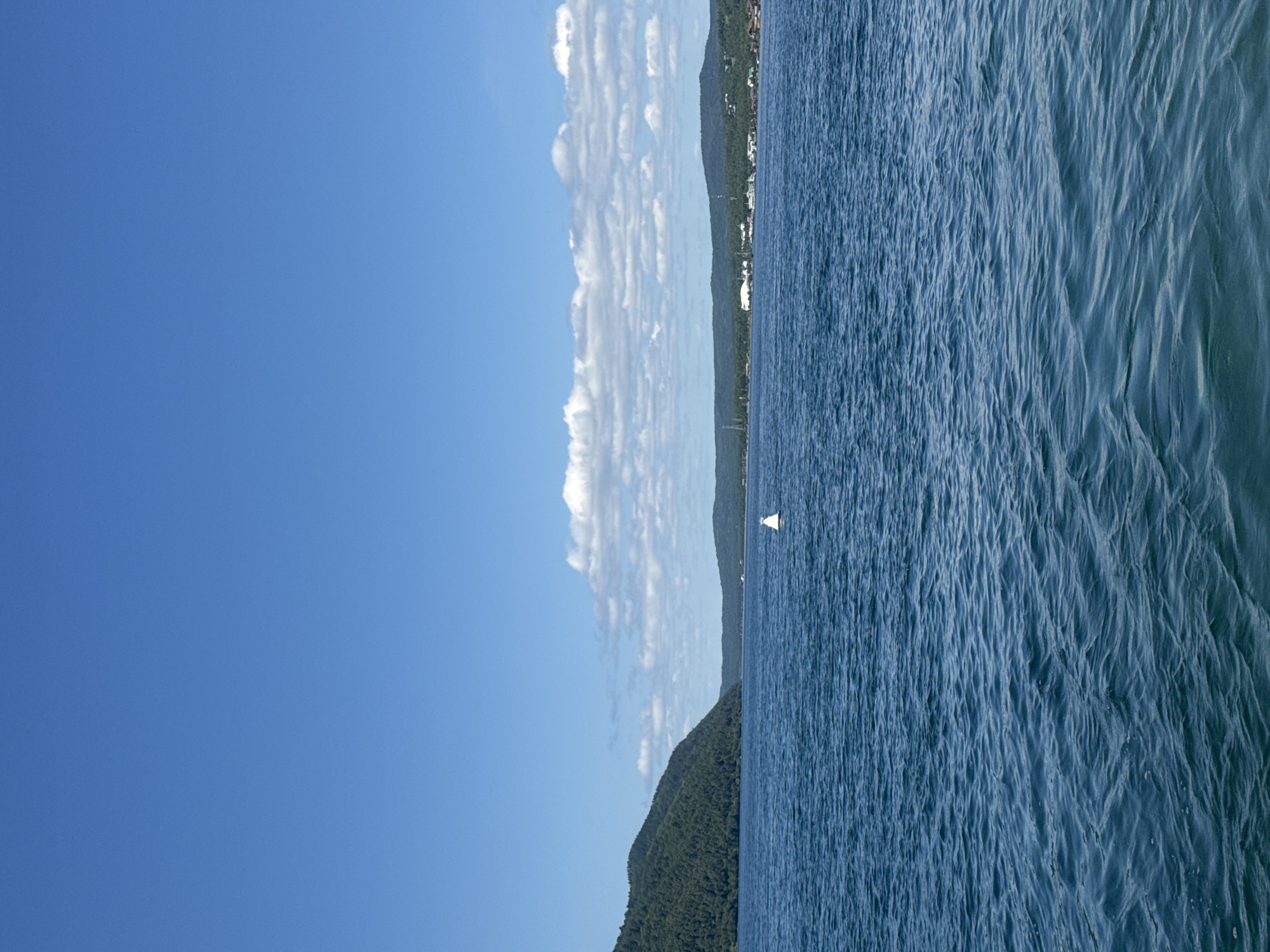 The image shows a wide expanse of deep blue water under a clear sky with scattered white clouds. On the left, a large, tree-covered hill slopes down to meet the water. In the distance, across the water, lies a shoreline with more tree-covered hills, and a line of buildings and infrastructure, including what appear to be industrial structures and possibly houses, is visible. A white buoy floats on the water in the middle ground. The scene suggests a natural landscape with some human development along the coast. The location is identified as Listvyanka, Russia.