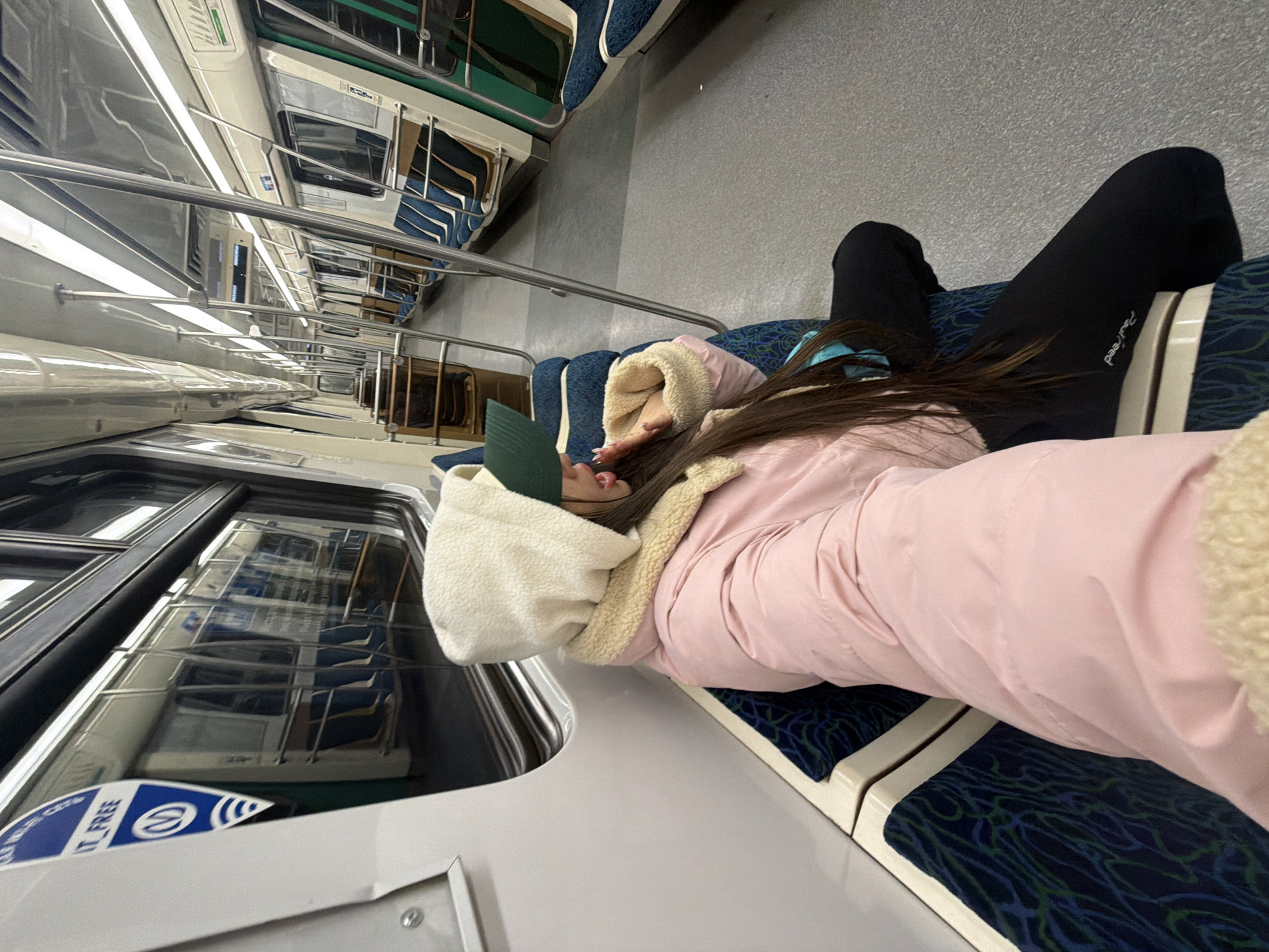 A person wearing black leggings, a pink jacket with a cream-colored hood, and a green cap is lying across two blue and green patterned seats on a train. The train's interior features overhead lights, handrails, and rows of forward-facing seats with blue and green upholstery. The train windows reveal reflections of other seats within the train car. A sticker on the train door is partially visible. The setting suggests an urban transportation context. The location is Saint Petersburg, Russia, based on the design of the train interior.