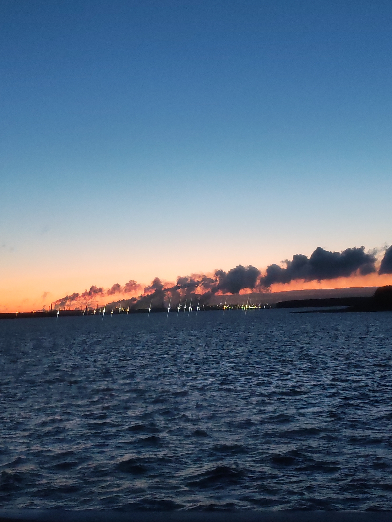 A wide shot captures a body of water under a sky transitioning from blue to orange at the horizon, with what appears to be a long series of industrial structures along the distant shoreline, emitting what could be smoke or steam. The water exhibits gentle waves, reflecting the dim light. The image suggests a sunset or sunrise scenario with a potential industrial presence in the background. The location is the city of Unknown, Unknown.