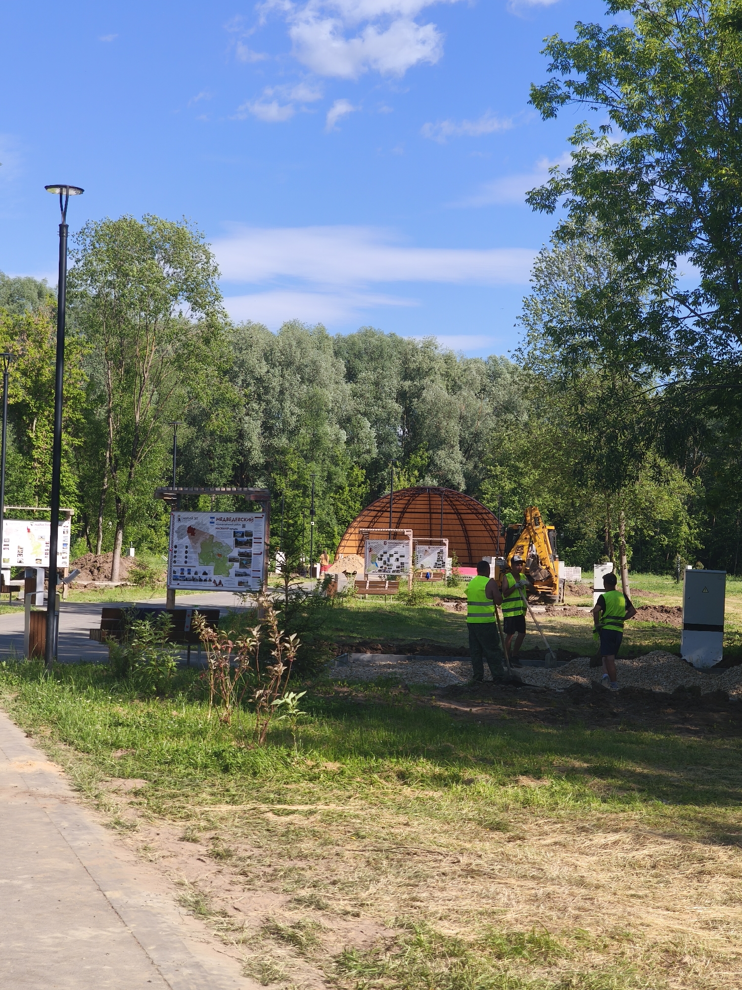 The image depicts an outdoor scene, likely in a park or recreational area, with a bright blue sky and scattered white clouds. In the foreground, a paved pathway runs along the left side, bordered by green grass and some dry straw. In the middle ground, there is a construction site where several individuals in yellow safety vests are working. One person is operating a yellow excavator, while others are digging with shovels. Behind the construction area, a large, dome-shaped wooden structure is under construction. Several informational signs are also visible, one of which appears to be a map. The background is filled with lush green trees. The scene suggests ongoing development or maintenance in a natural setting. The presence of informational signs with Russian text and the specific architectural style of the informational displays strongly suggest the location is the "Stone Flower" Ethno-Park in Yoshkar-Ola, Russia. The park is known for its themed zones, and the dome structure resembles elements often found in such cultural or recreational facilities.