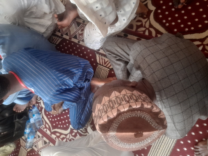 The image shows four men sitting on a red and white patterned carpet in Gusau, Nigeria. The man on the left, wearing a grey and white checkered tunic, is sitting on the floor with his back to the viewer. He is wearing a brown and beige cap with a decorative pattern. The man in the center is wearing a blue striped tunic and is sitting cross-legged with his legs bent. The man to the right is wearing a white tunic. He is sitting with his legs crossed, and his right leg is bent at the knee. The fourth man is partially visible, wearing a white tunic with a red sash. He is sitting on the floor with his legs extended and is leaning back on the man in the blue striped tunic. Several plastic water bottles are visible in the background. The location of the image is an indoor space with a carpeted floor, possibly a mosque.