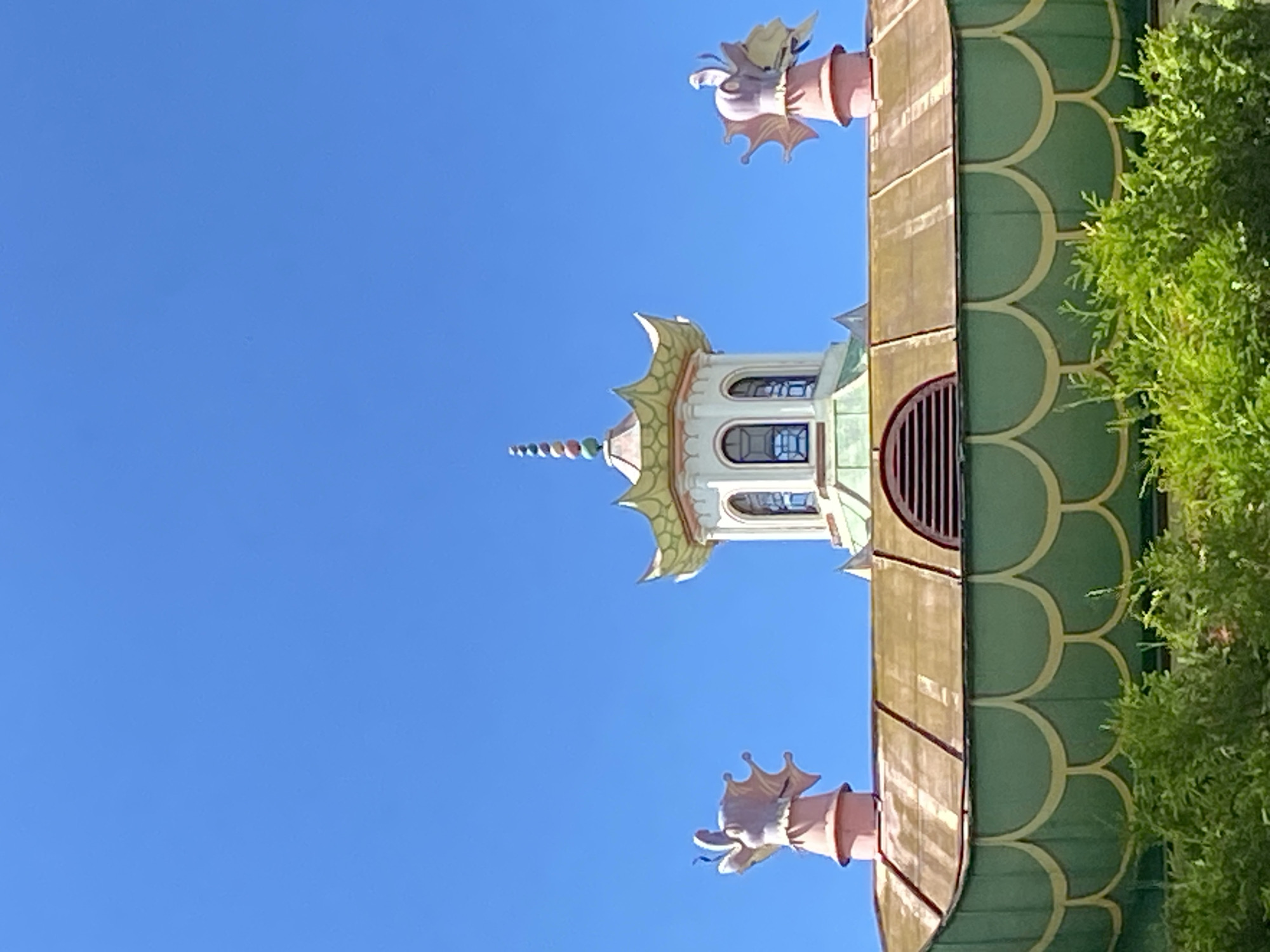 The image shows the roof of a building with distinct architectural features, likely in the city of Pushkin, Russia, given the context of the prompt. The building's roof is a prominent element, featuring a multi-tiered design with a central octagonal turret topped by a spire composed of colorful, stacked spheres. Flanking the central turret are two smaller, pink structures, each adorned with decorative dragon-like figures. The roof itself appears to be constructed of copper or a similar metallic material, exhibiting a golden-brown hue. Below the main roofline, a green, scalloped facade is visible, accented with yellow trim, and a semicircular vent with horizontal slats is present on the upper portion of the main roof. Lush green foliage, possibly trees or large bushes, partially obscures the lower section of the building. The sky above is a clear, vibrant blue, indicating fair weather conditions. There are no people visible in the image.