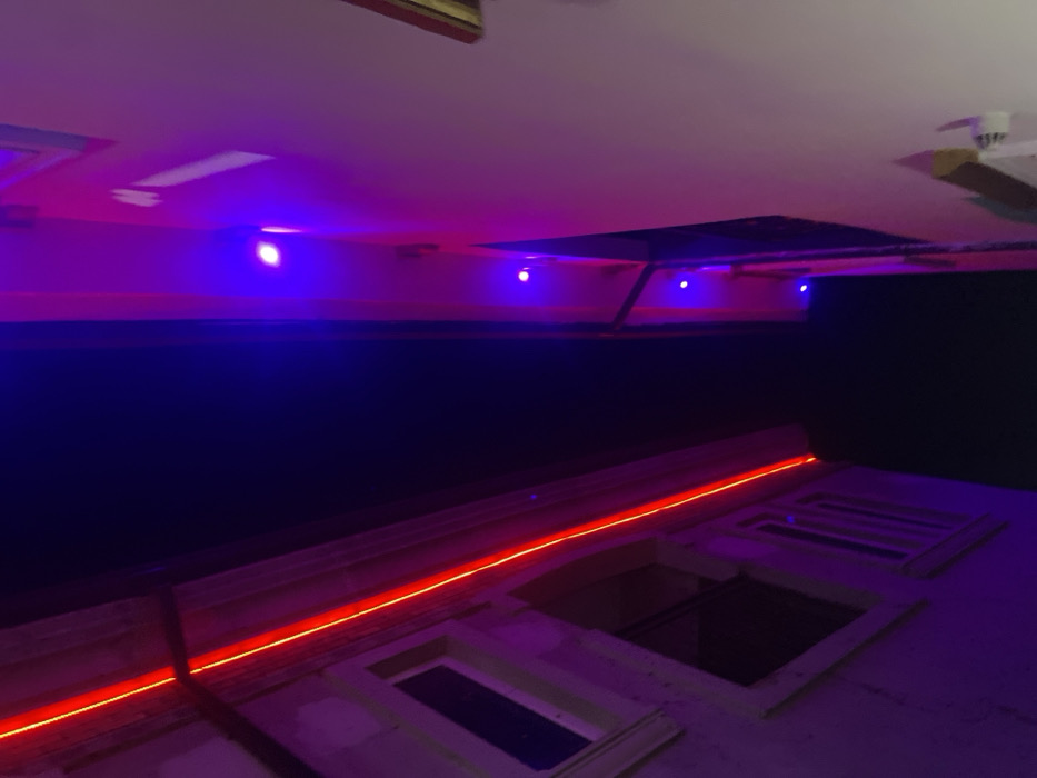 The upside-down image shows a section of a building exterior at night. A bright orange light strip highlights the edge of the building and window frames. Above, a dark wall extends to the ceiling. The ceiling has recessed lighting and blue LED strip lights. A partial view of a ceiling fan is visible. The color scheme is dominated by hues of purple, blue, and orange.