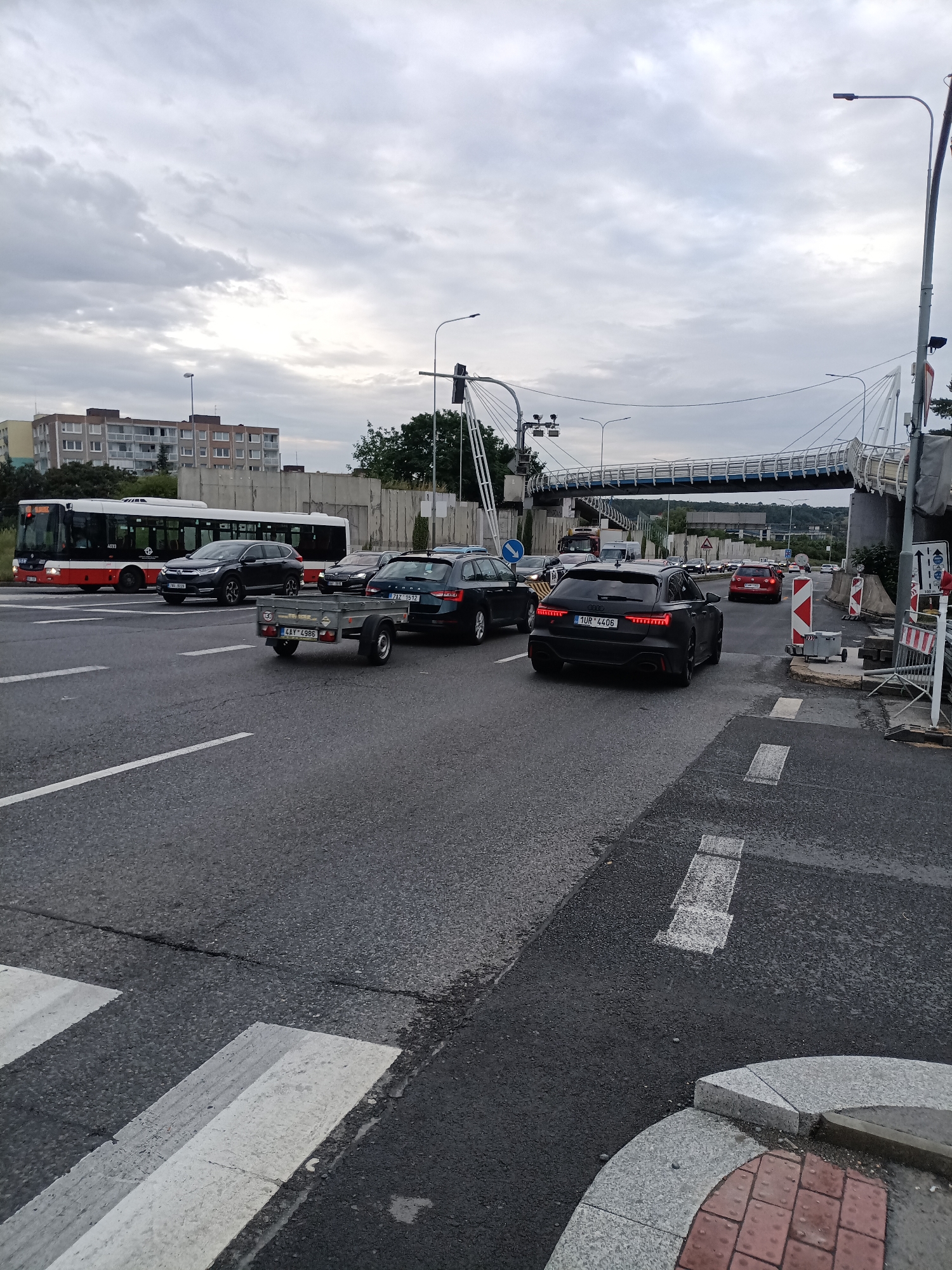 This media file captures a street scene in Prague, Czechia, during the daytime under an overcast sky. The image focuses on a multi-lane road with several vehicles, including a bus, cars, and a trailer.
In the foreground, a crosswalk is visible, with a small, grey trailer being towed by an unseen vehicle. To the left, a red and white public bus is stopped, and next to it, a dark grey SUV. Further down the road, another dark grey sedan and a black Audi are prominent. The black Audi, with license plate "1UR 4406", is facing away from the camera.
In the background, a modern pedestrian bridge spans across the road. Buildings can be seen on the left, and trees and more traffic are visible further down the road. Construction barriers and signs indicate some ongoing work or road closure. The overall mood is neutral, with no discernible emotions. The time of day appears to be late afternoon or early evening, given the cloudy conditions. No people are clearly visible in the scene.