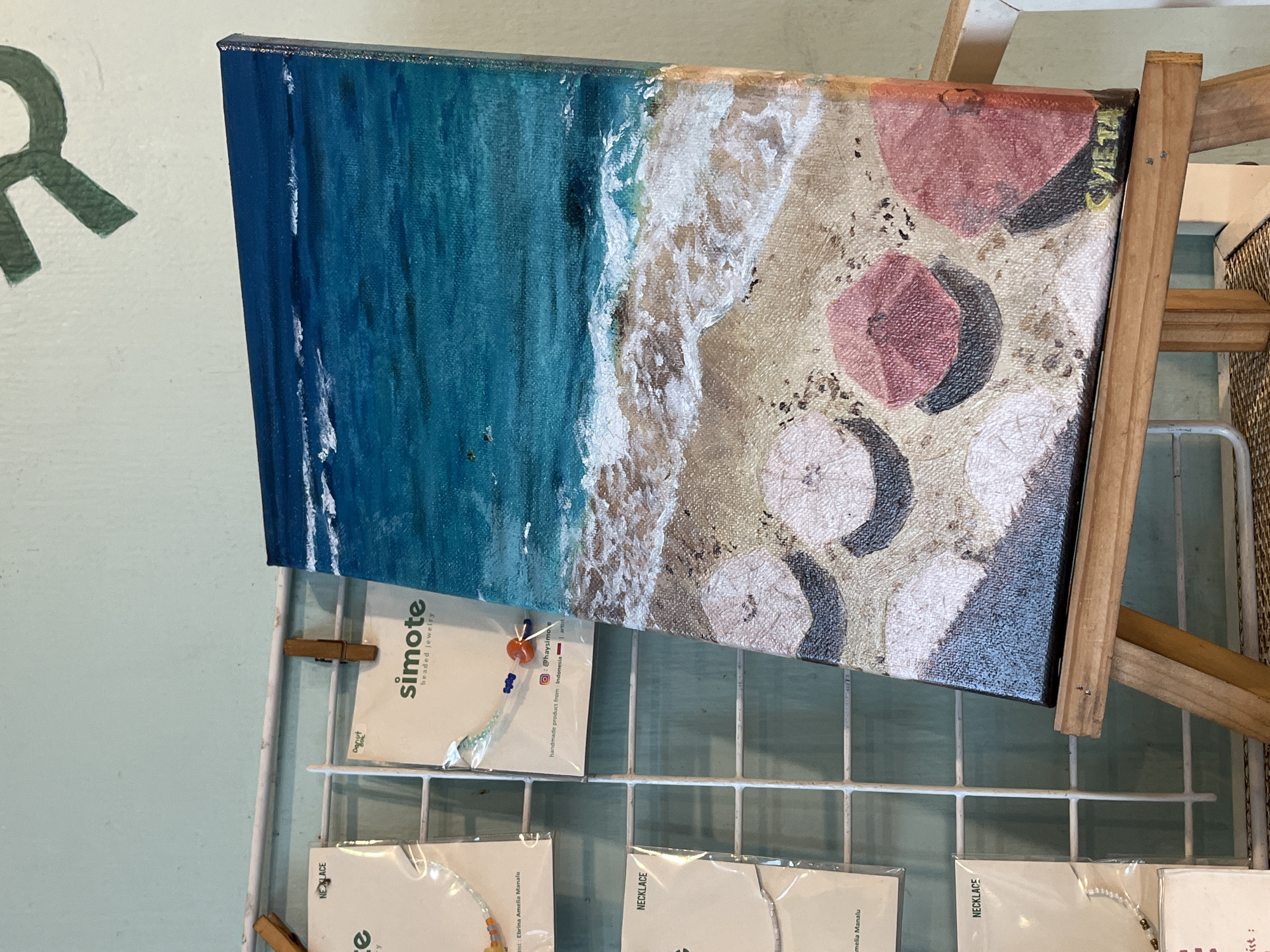 The image shows a painting on display in a retail setting. The painting depicts a beach scene with blue water, white waves, and sand with pink umbrellas. The painting is propped up on a small wooden easel, which is placed on a white wire grid. Several cards with the brand name "simote beaded jewelry" are clipped to the grid below the painting. The cards indicate the jewelry is a handmade product from Indonesia and refers to @haysimove on Instagram. One card is labeled "Danut," and others are labeled "NECKLACE" with the artist's name listed as "Ebrina Amelia Manalu" or "amelia Manalu". The environment is a retail space where the jewelry and painting are likely being sold, however, location in the city of Unknown, Indonesia is unable to be deciphered. There are no people visible in the image.