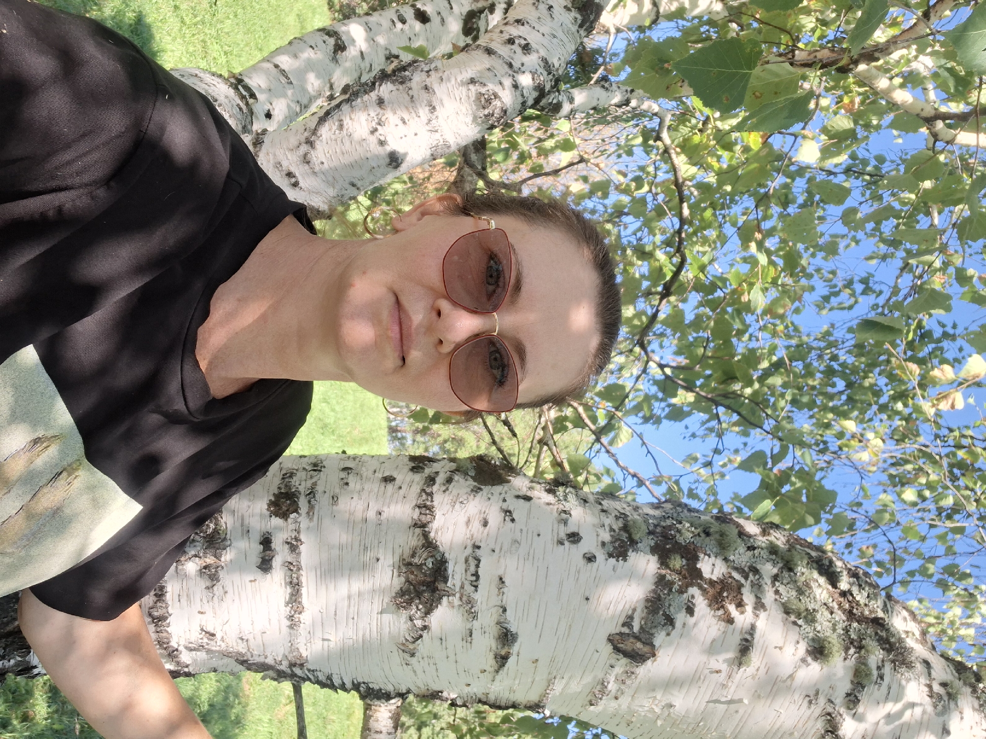A woman wearing sunglasses and a black t-shirt stands outdoors, leaning against a white birch tree. The tree has distinctive dark markings on its bark. Green leaves and branches of the tree are visible, as well as a blue sky with some clouds. The ground is covered in green grass. The scene appears to be located in Altaiskoe, Russia.