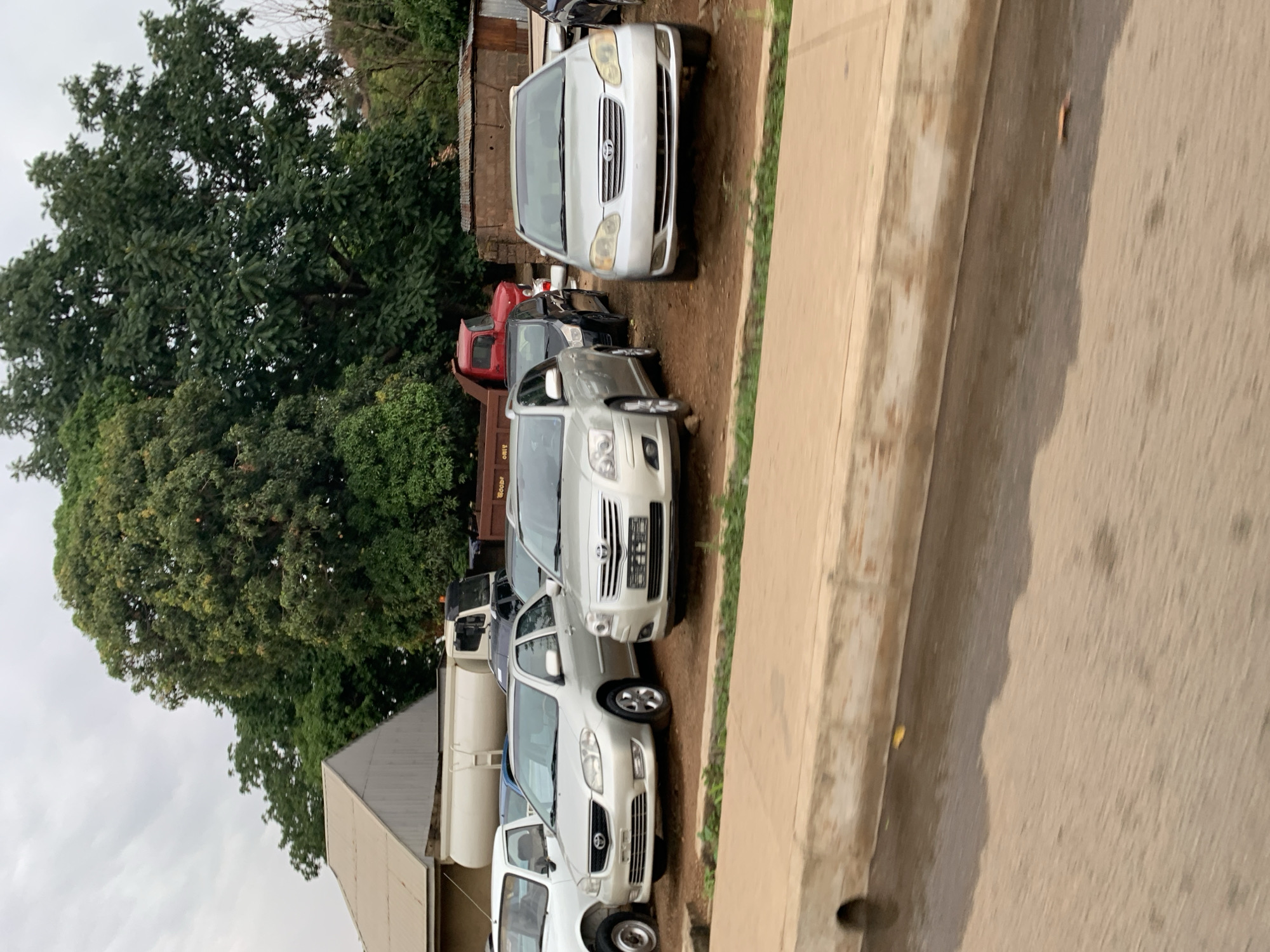 The image shows a row of cars parked along a street in New Stadium, Nigeria. The cars are various models, primarily white and silver sedans. To the left of the vehicles is a large tree and a building with a white wall and a corrugated metal roof. The street is paved and appears to be wet in some areas. The sky is overcast. There are no visible people. A red truck is seen behind the first silver sedan.