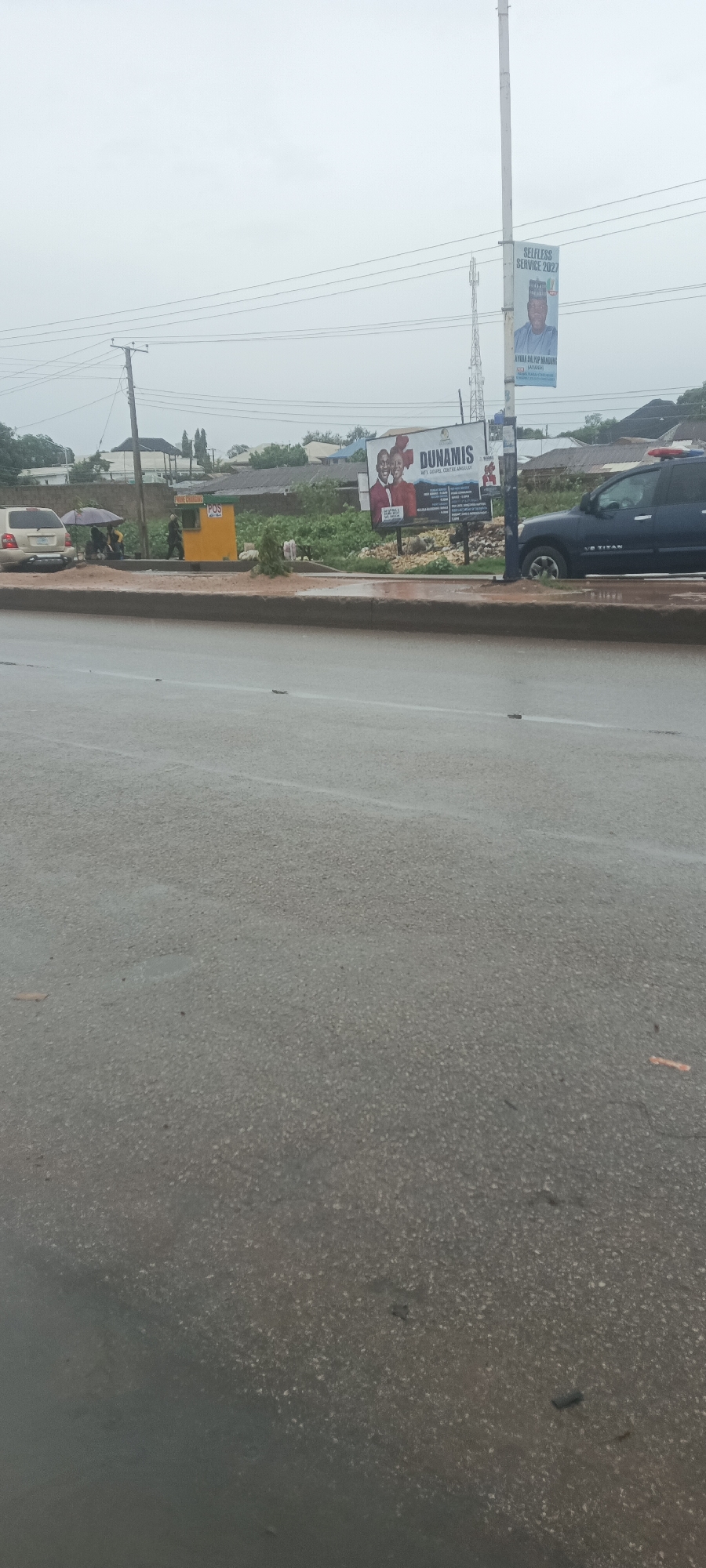 The image depicts a street scene in Bukuru, Nigeria, under an overcast sky, suggesting a dull day or recent rainfall, as indicated by the wet asphalt road. A concrete median separates the visible lane from the opposite direction.
On the left side of the road, several people are gathered on a shoulder or sidewalk. One person stands under a dark umbrella, while another is near a yellow kiosk labeled "PURE CHANNEL" and "POS." A light-colored SUV, possibly a Toyota Highlander, is parked nearby. In the background, there are various buildings and green vegetation.
Further along the street, a large billboard prominently displays "DUNAMIS INT'L GOSPEL CENTRE," with visible text for prayer points and service times. Below this billboard, piles of what appear to be corn cobs are visible on the ground.
On the right side of the road, a dark-colored SUV, specifically a Nissan Titan, is partially visible. A tall utility pole stands beside the road, from which hangs a political banner. This banner features a man in traditional attire and includes the text "SELFLESS SERVICE 2027," along with parts of a name "AYUBA BAL/POP MANGUNS" and "JOS SOUTH." Power lines traverse the upper portion of the frame. The overall scene captures daily life in a Nigerian town setting.