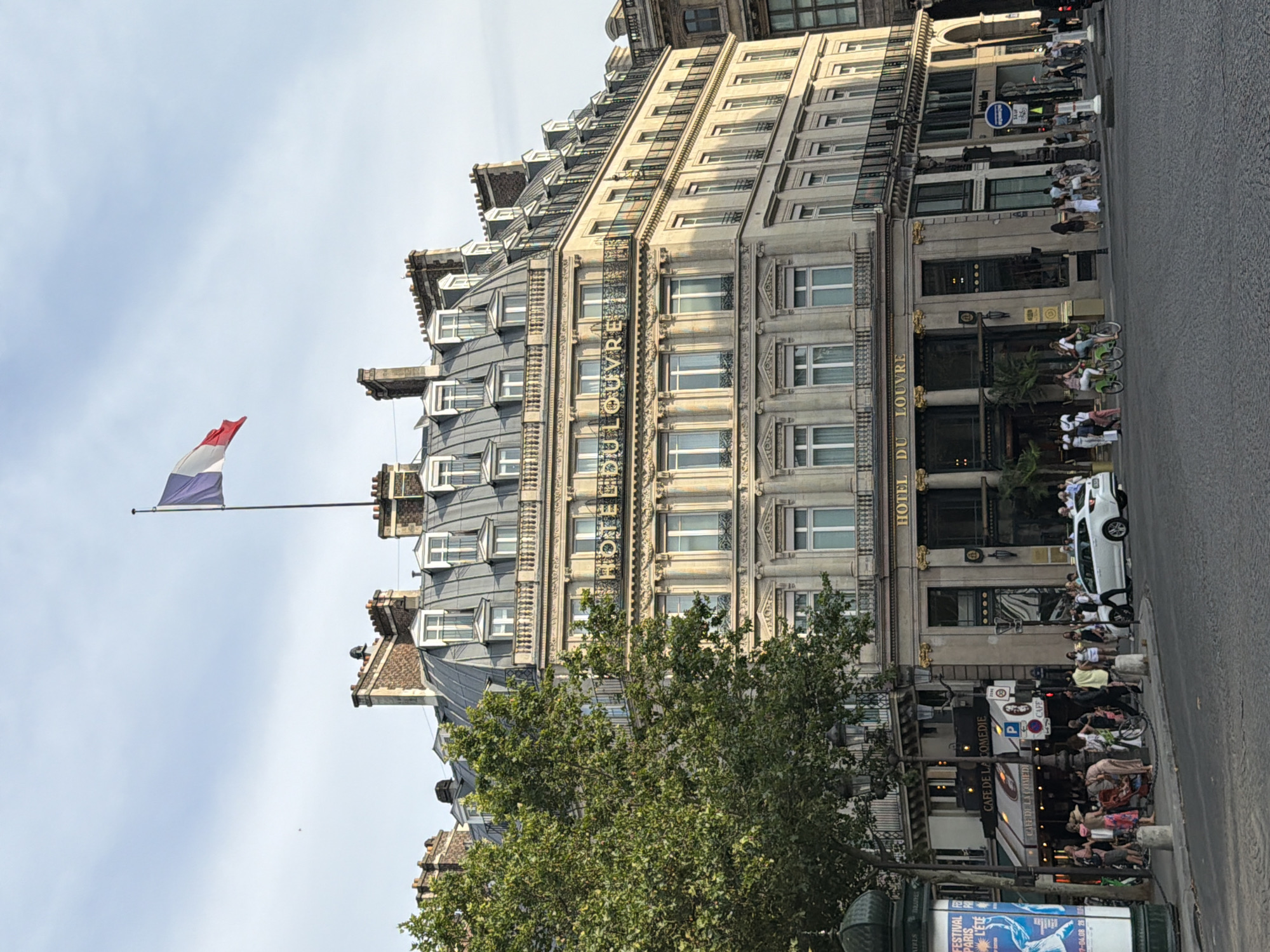 The image shows the exterior of the Hotel du Louvre in Paris, France, under a clear sky. The hotel is a large, ornate building with multiple stories, featuring a mansard roof and classical architectural details. A French flag flies from a flagpole on top of the building. In front of the hotel, a busy street scene unfolds. People are walking on the sidewalk, some are seated at outdoor cafe tables, and others are cycling. A white car is parked on the street, and a tall, green tree stands to the left of the building. The scene is illuminated by natural daylight, casting shadows on the building and street.