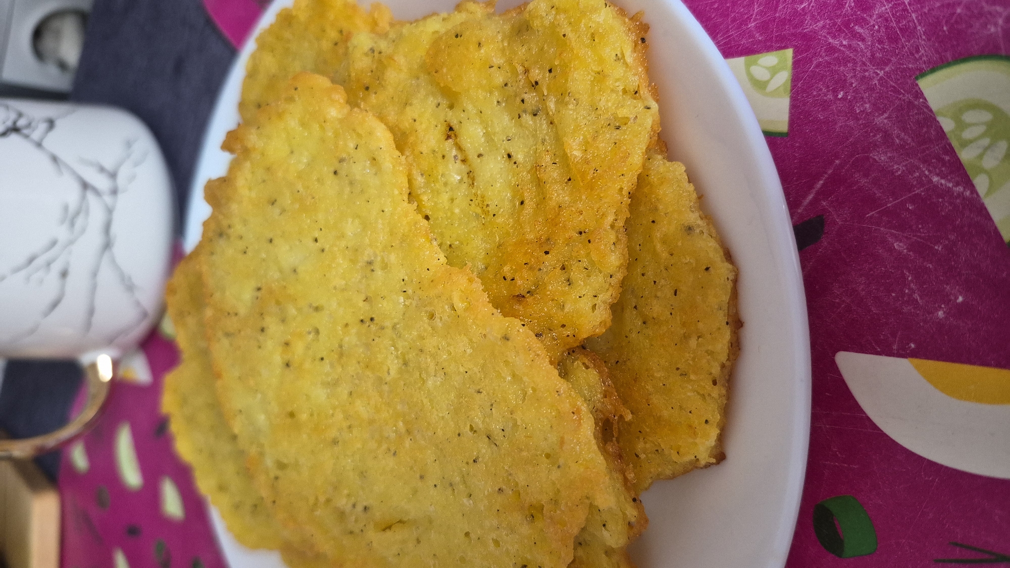 A close-up, slightly overhead shot shows a stack of golden-yellow, thin, crispy potato pancakes on a white plate. The pancakes are irregular in shape with slightly ruffled edges, and speckled with black pepper. They are piled on top of each other, with the bottom layers partially obscured by the top ones. In the background, out of focus, is a white mug with a marble-like pattern and a gold handle. The plate rests on a vibrant pink surface decorated with illustrations of sliced cucumbers and green onion rings. The overall lighting is warm, highlighting the texture and crispness of the pancakes. The location is not specified in the image.