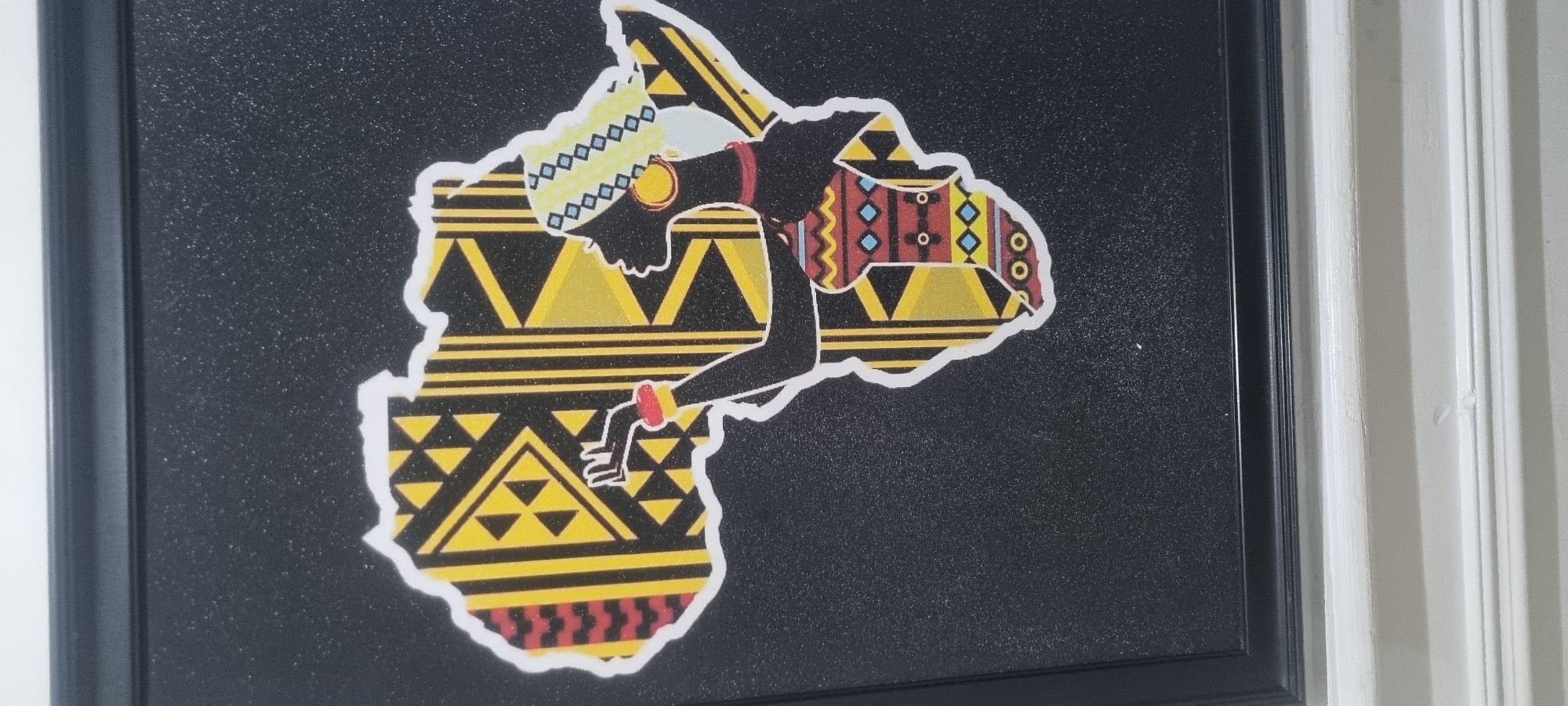 A framed artwork featuring a stylized depiction of the African continent with a woman's silhouette positioned within its borders is mounted on a wall. The artwork is characterized by a black background, over which a yellow and black patterned image of the continent is overlaid. The silhouette of the woman is black, adorned with colorful geometric patterns suggesting traditional African attire and jewelry. The artwork is framed in a black frame. The scene is located indoors, adjacent to a white wall. There is no information on the location of the city of Alasia, Nigeria.