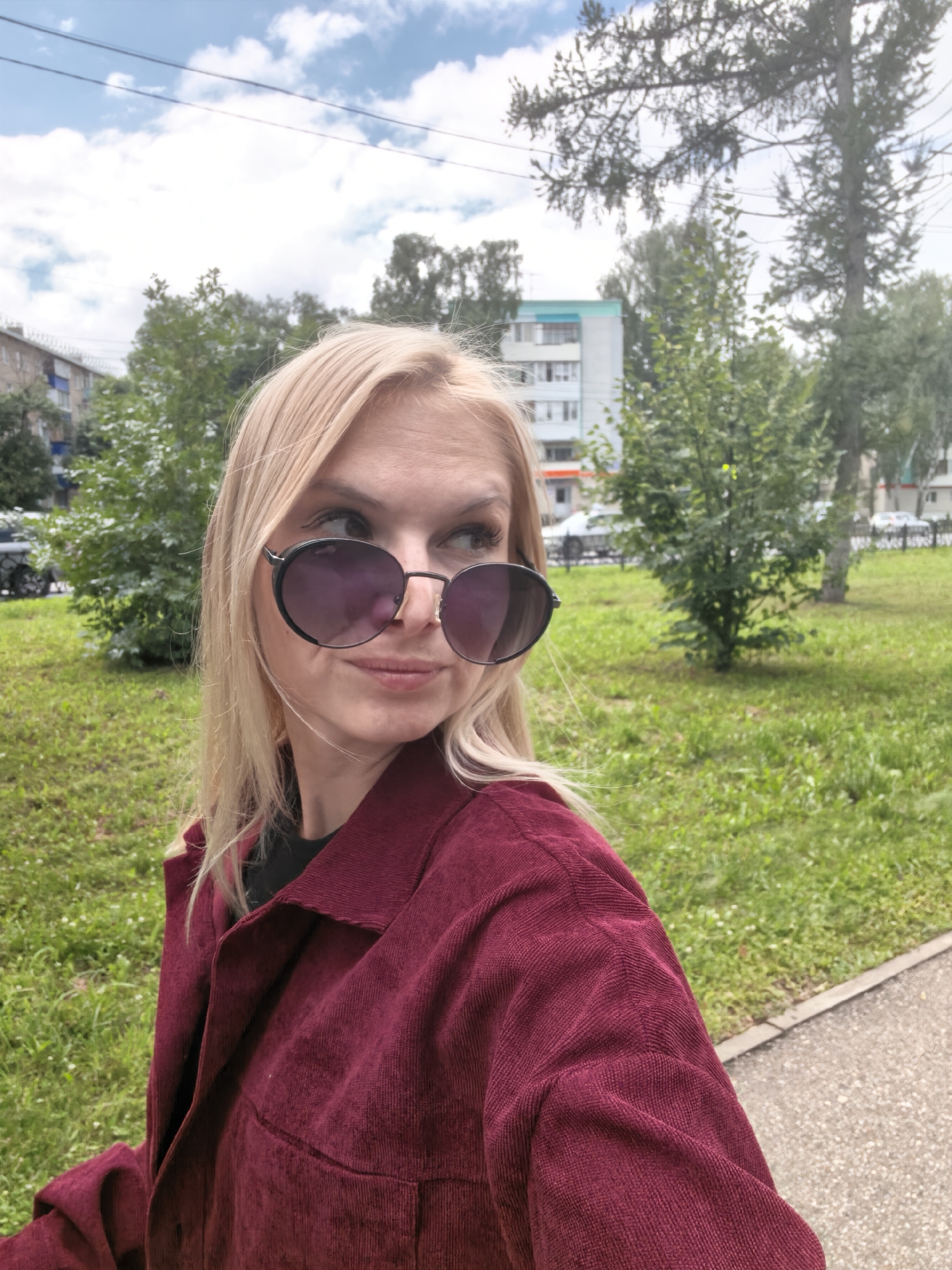 In a medium shot, a blonde woman wearing large, dark-lensed sunglasses and a burgundy corduroy jacket stands outdoors. She is looking to her right, away from the camera, with a slight smile. The background suggests an urban environment. A sidewalk is visible in the foreground to the right, bordered by a grassy area. Behind the woman, there are green trees, a multi-story apartment building, and more trees. The sky above is partly cloudy. The scene appears to be in the city of Sterlitamak, Russia, as indicated by the architectural style of the buildings and the general appearance of the urban greenery.