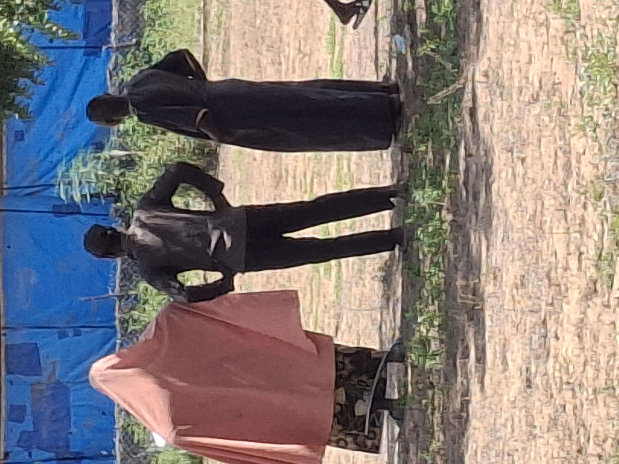 Three individuals are standing outdoors in Batungo, Nigeria, with their backs to the camera. The person on the left is covered by a large, light pink garment, with a patterned skirt visible underneath. The middle person is wearing a dark, collared jacket over dark pants. The person on the right is wearing a long, dark robe. Behind them, a large blue tarp is stretched taut, with some greenery and trees visible. The ground is a mixture of dirt and sparse grass. The lighting suggests it is daytime.