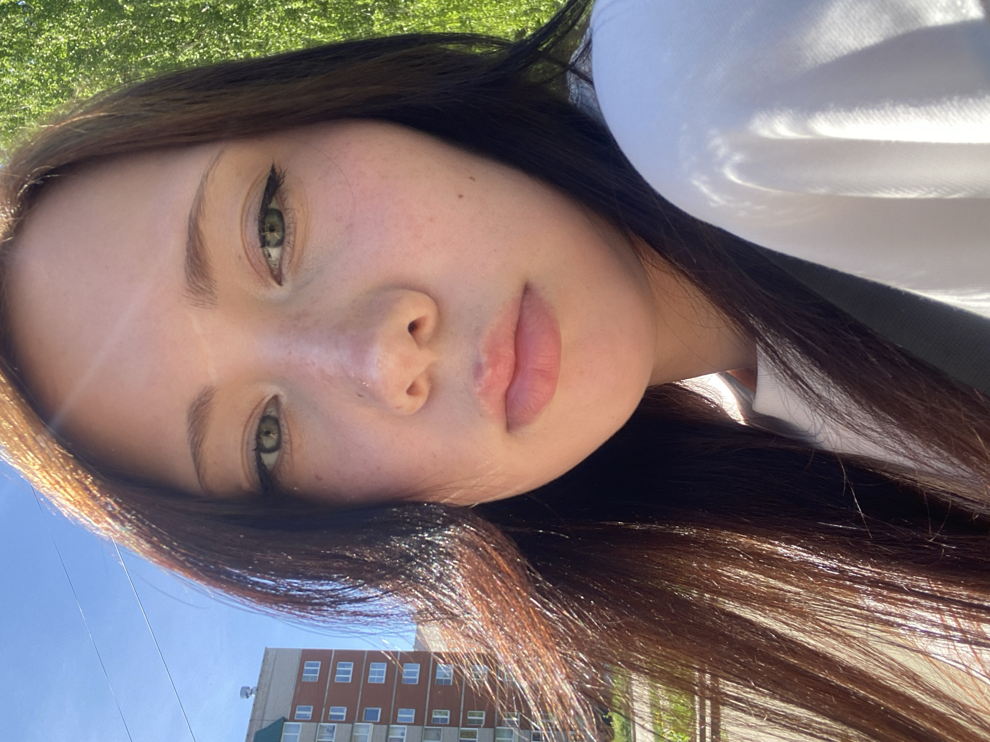 A close-up, angled selfie of a young woman with light skin, dark brown hair, and green eyes. She has visible eyelashes, and light makeup on her eyes. Her lips are full and light pink in color. The background includes glimpses of a building with multiple windows, a blue sky, and green vegetation. The scene appears to be outdoors in an urban or suburban setting.