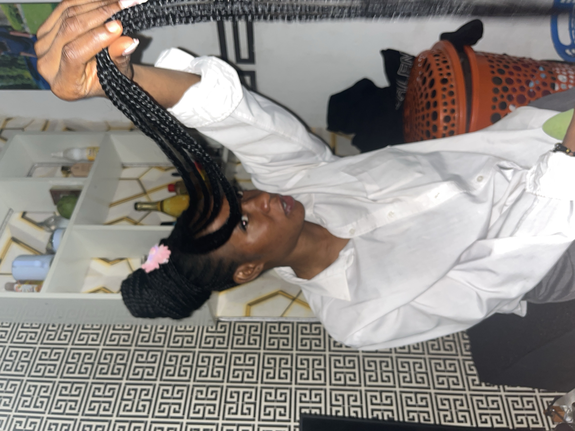The media file features a young Black person in a brightly lit indoor setting, likely a home. The person is wearing a white collared shirt and is in the process of braiding or styling their hair. Their hands are actively manipulating black braided extensions. A pink flower accessory is visible in their hair. The background includes shelves with various items, possibly toiletries or decorative objects, and a distinctive black and white geometric patterned floor. To the right, an orange basket with circular cutouts is partially visible, along with some dark clothing. The scene appears to be during the daytime due to the ample lighting. The overall impression is one of personal grooming or preparation.