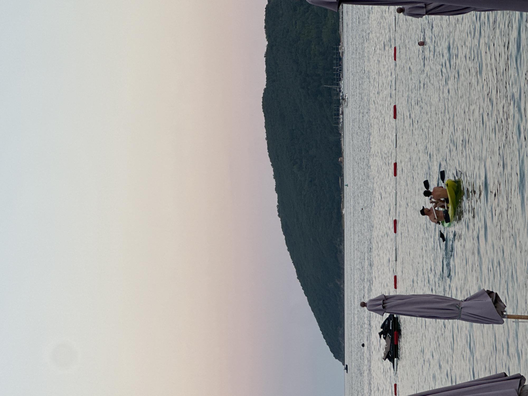 The image depicts a serene scene at the coast of Budva, Montenegro, likely during the golden hour as suggested by the soft, gradient sky transitioning from a pale blue to a gentle pink. In the foreground, the calm, rippling waters of the Adriatic Sea are visible, dotted with beach umbrellas. Two individuals are seated in a yellow kayak, paddling together. Further out in the water, a jet ski is parked near a floating line of buoys, and a person can be seen swimming. In the mid-ground, a densely forested green hill slopes down towards the sea, with a few boats anchored in the distance near the shore. The overall impression is one of tranquility and leisure.