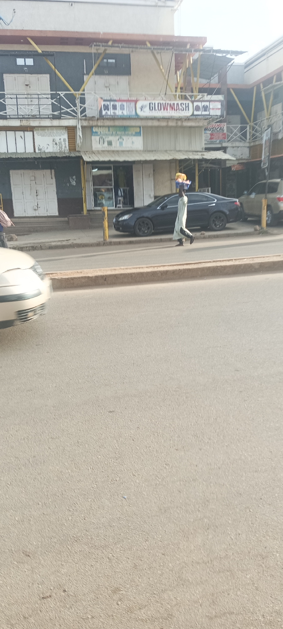 The image shows a street scene in Jos, Nigeria. A person is walking along the sidewalk carrying items on their head. Several cars are parked on the side of the road. Buildings line the street, with shops and businesses visible, including "GLOWMASH" and "OKASON RECORDING LIMITED". The road appears to be paved. The sky is overcast.