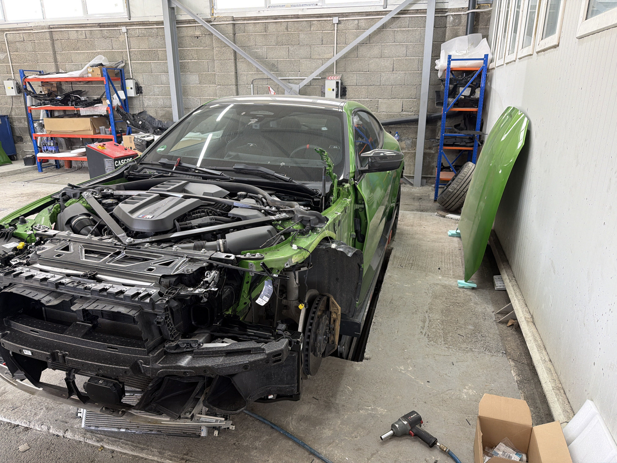 The image captures a partially disassembled, bright green sports car in what appears to be an automotive workshop or garage. The car, likely a BMW M-series judging by the "BMW M Power" inscription on its exposed engine cover, has its front bumper, grill, and possibly front fenders removed, revealing the radiator, intercooler, and other engine bay components. The car's hood is detached and leans against a light-colored wall on the right side of the frame. The front driver-side wheel well is also exposed, showing the brake disc and caliper, and the suspension components.
The setting is an indoor space with a concrete floor and block-patterned concrete walls. High on one wall, there are several rectangular windows, suggesting natural light, supplemented by artificial lighting. In the background, two blue metal shelving units with orange shelves hold various car parts, including what look like bumpers, trim pieces, and cardboard boxes. A red "CASCO" battery booster or charger sits on one of these shelves. Another single tire is visible near the wall. On the floor in the foreground, an impact wrench with an air hose attached, bearing the "WURTH" brand name, and an open cardboard box are visible, alongside a detached radiator or intercooler unit. No people are visible in the scene. The overall impression is one of active repair, maintenance, or customization.