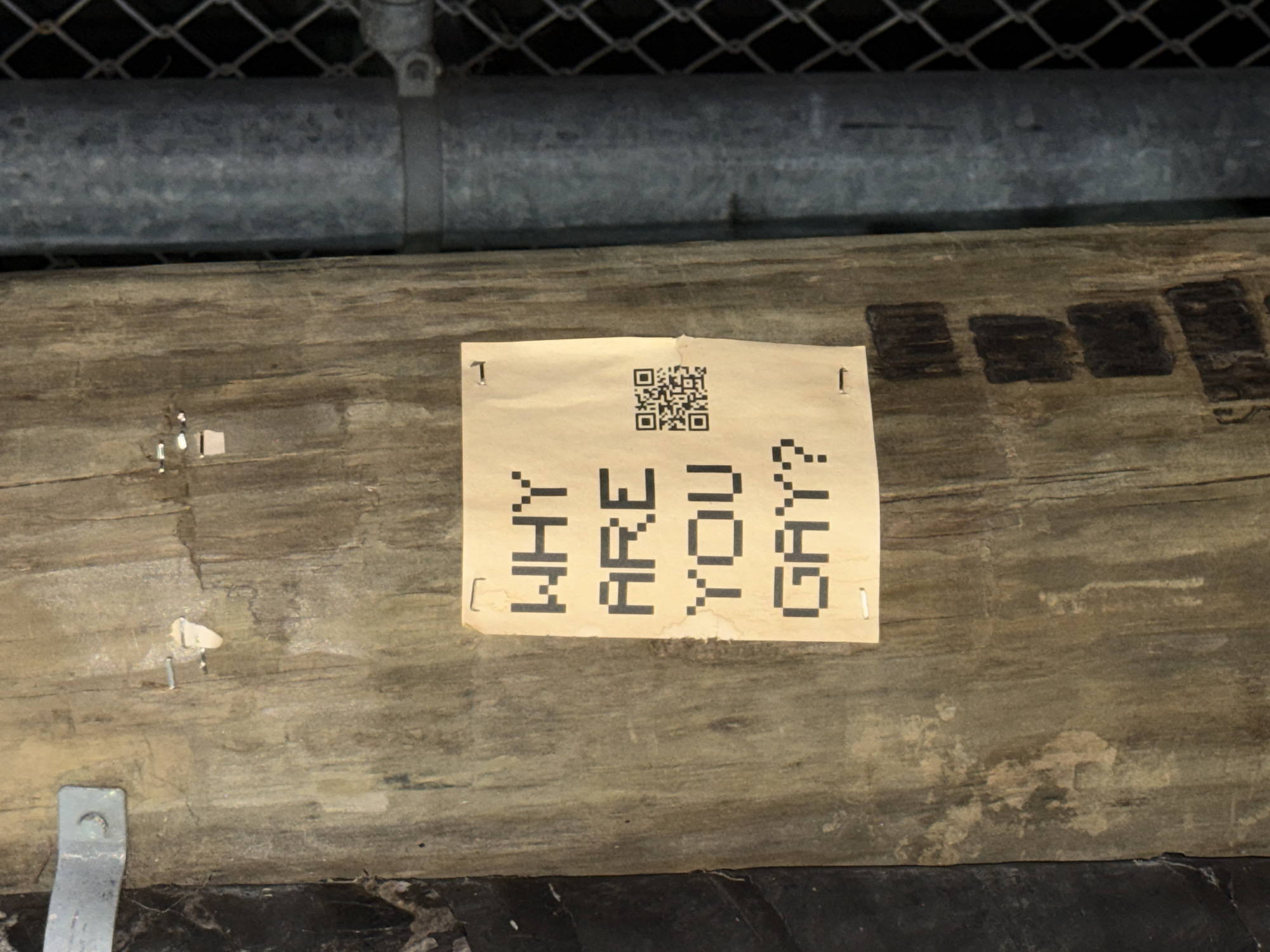 A piece of light brown paper is stapled to a dark, weathered wooden beam. The paper features large, black, block-lettered text arranged vertically, which reads "WHY ARE YOU GAY?". Above this text, a black QR code is prominently displayed. Staples are visible securing the paper to the wood.
The wooden beam, textured with grain and showing signs of age and wear, runs horizontally across the midground of the image. Above the beam, a horizontal metal pipe or conduit is visible, secured by a clamp. Behind the pipe and extending upwards, a chain-link fence is discernible, suggesting an outdoor or semi-enclosed public space. The overall lighting is dim, casting a somewhat muted tone over the scene. The image is set in West Hollywood, United States of America. No people are visible, and no activity is taking place.