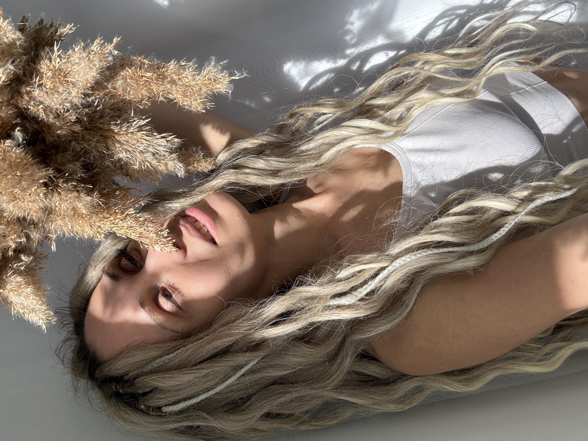 An overhead medium shot captures a person with long, wavy blonde hair lying on a light-colored, possibly white, surface. The individual is positioned diagonally across the frame, with their head towards the upper left. They are wearing a simple white ribbed tank top. Their eyes are partially closed or looking downwards, and their lips are slightly parted, suggesting a relaxed or contemplative state.
A prominent bunch of dried, light brown ornamental grass or reeds, likely pampas grass, occupies the upper left quadrant of the image, partially covering the person's face on that side. The person's long, flowing blonde hair, which appears to have integrated thin white braided strands, cascades around their head and shoulders, spreading across the surface.
The scene is illuminated by strong, direct sunlight, casting distinct, sharp shadows across the surface and the person's form. These shadows, some resembling abstract organic shapes or foliage, create a high-contrast visual effect. The lighting emphasizes the textures of the hair, the dried grass, and the ribbed fabric of the tank top. No visible text is present in the image. The location context for the image is Rusanovo, Russia.