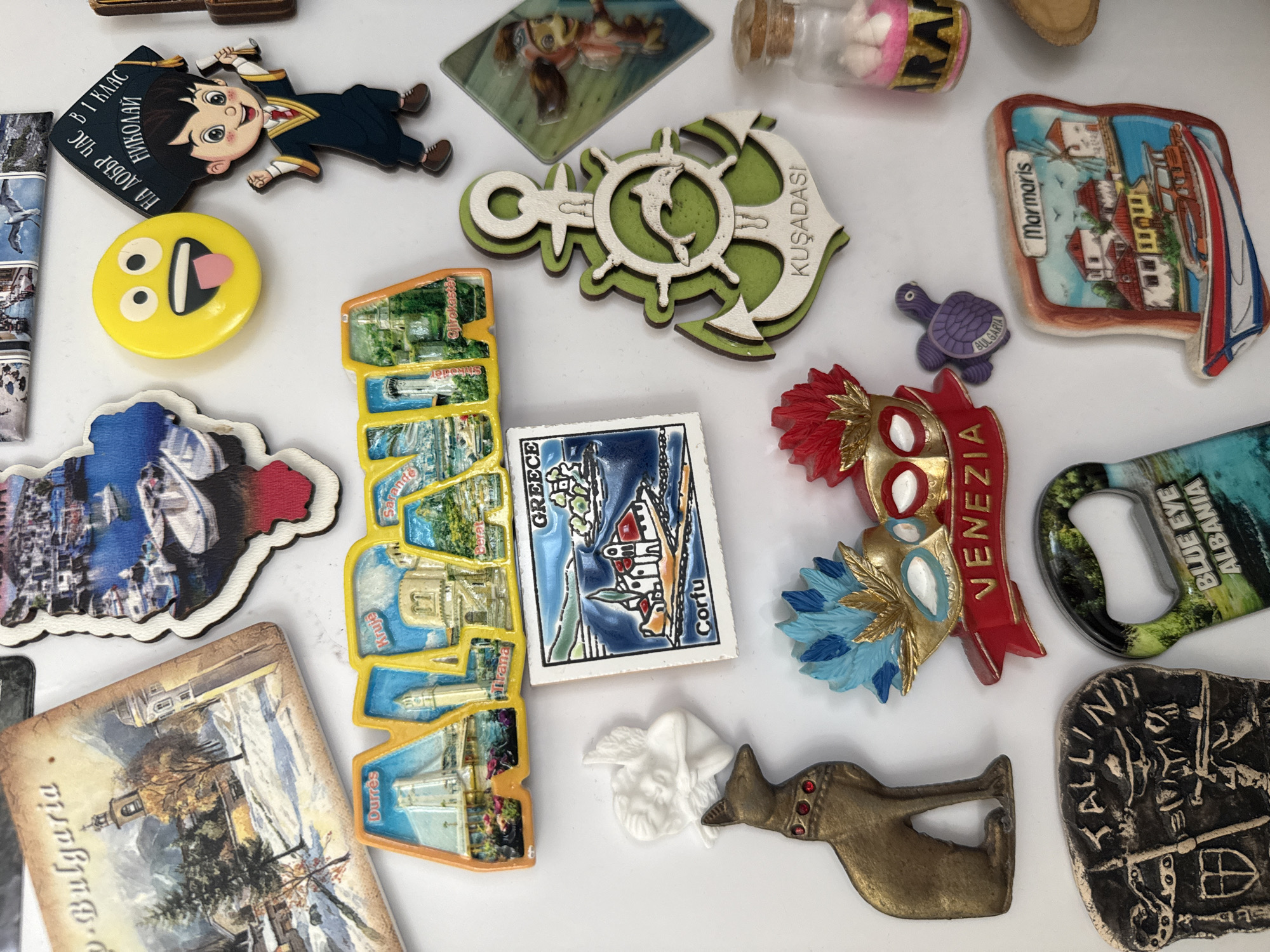 The image shows a collection of souvenir magnets from various locations. Magnets from Bulgaria (including a postcard-style magnet and a turtle with "BULGARIA"), Albania ("BLUE EYE ALBANIA" and a tiered magnet), Greece (Corfu, and another tiered magnet displaying "Durres", "Kruje", "Sarande", and "Gjirokaster"), Turkey (Kuşadası, Marmaris) and Italy (Venezia) are present. There is a cartoon character wearing a graduation gown with the Bulgarian text "НА ДОБЪР ЧАС В 1 КЛАС НИКОЛАЙ". There is also a yellow smiley face. The magnets are laid out on a white surface. The city of Elhovo, Bulgaria is not mentioned in the image.