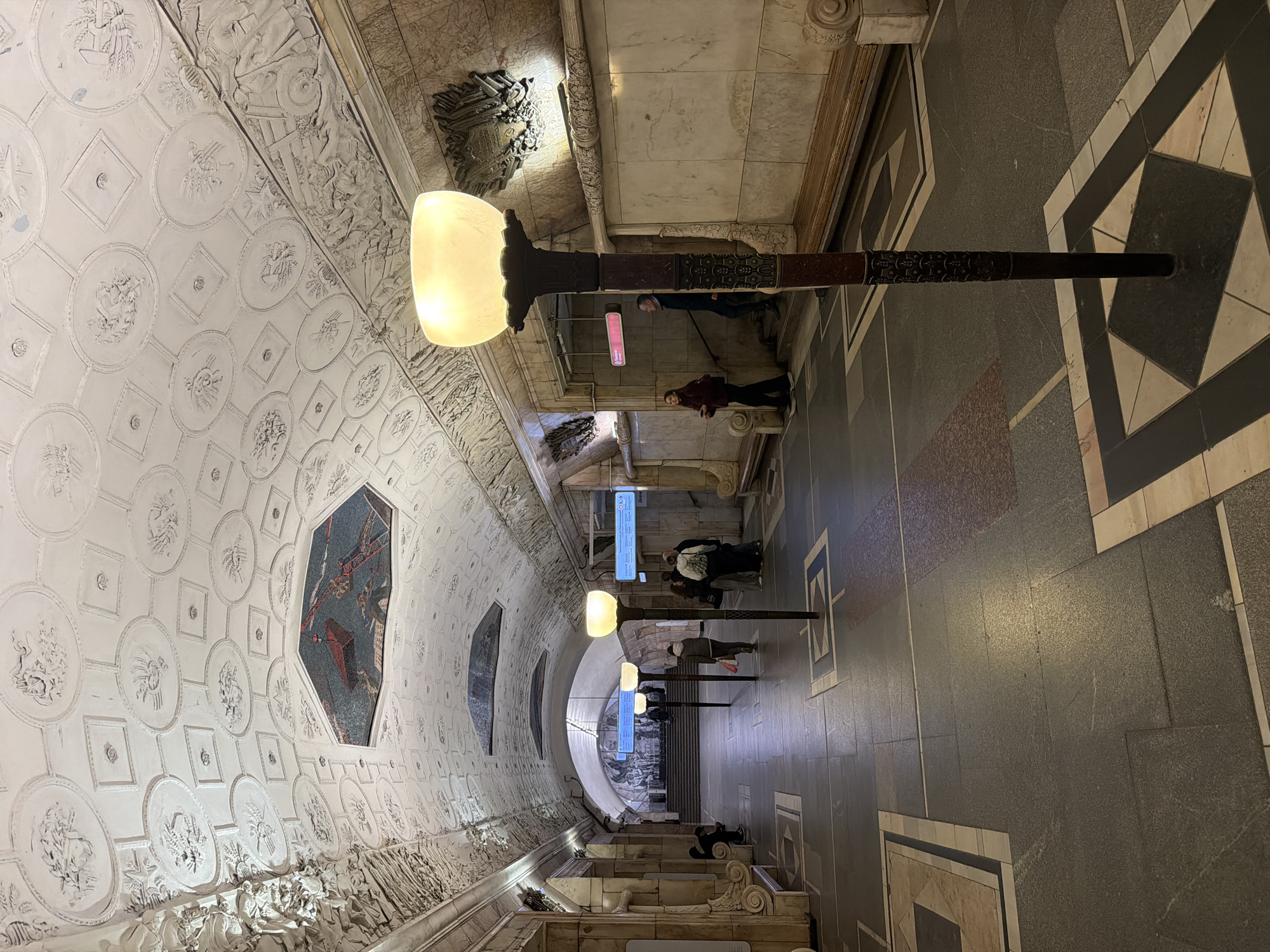 This media file captures a grand and ornate underground passage, likely a station hall or entrance within the Moscow Metro. The perspective looks down a long, wide corridor that appears to descend, possibly towards an escalator well or further platforms.
The left side of the passage is dominated by a highly decorated vaulted ceiling or upper wall, painted in white. This surface is covered with intricate bas-reliefs, featuring a grid of circular and square panels. The circular panels depict various motifs, including what appear to be agricultural elements like wheat stalks and flowers, alongside other symbolic figures. Below these, a continuous frieze of detailed bas-reliefs stretches along the wall, portraying numerous human figures engaged in different activities, possibly a celebratory procession or scenes of labor. Several large, colorful mosaic panels are integrated into this decorative surface, one prominently showing figures and abstract shapes in shades of blue, red, and gray.
Along the right side of the passage, several tall, dark-colored floor lamps with large, glowing, light-yellow diffusers provide warm illumination. The floor itself is paved with large geometric patterns, created using dark gray, black, white, and light-colored marble or stone tiles.
In the mid-ground and background, several people are visible, mostly standing or walking further into the hall. They appear to be commuters or visitors, some dressed in winter coats. Above them, multiple blue rectangular signs or information screens are suspended; the text on these is largely unreadable due to distance, but some Cyrillic characters are faintly discernible on a closer screen.
The scene conveys a sense of historical grandeur and artistic opulence, characteristic of the Moscow Metro's famous architecture. The time of day is not apparent due to the indoor setting and artificial lighting.