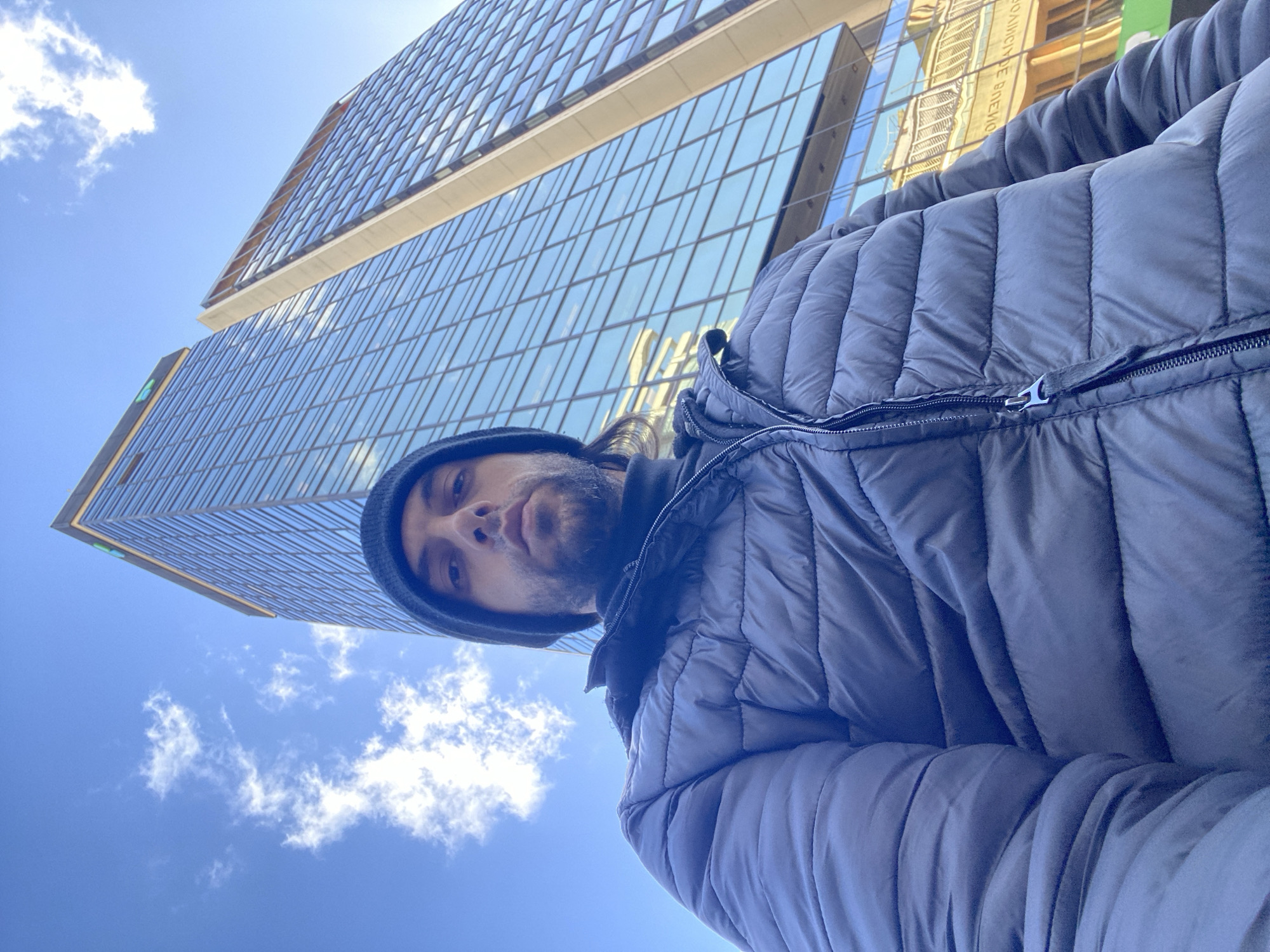 The image is a low-angle selfie featuring a man in the foreground, against a backdrop of a modern glass skyscraper and a bright blue sky. The man, seen from the chest up, has a dark beard and wears a dark beanie and a dark, padded puffer jacket with a visible zipper pull. He looks directly at the camera with a neutral expression.
Behind him, a tall, contemporary building with a blue-tinted glass facade reflects its surroundings. The architecture suggests a modern urban environment. The sky is clear and bright blue with a few scattered white clouds, indicating it is daytime, likely morning or early afternoon, with sunny weather.
Faintly visible in the reflection on the building's glass surface is inverted, unreadable text. A small, indistinct green logo or sign is also present on a lower section of the building. The scene captures an urban setting in Buenos Aires, Argentina.