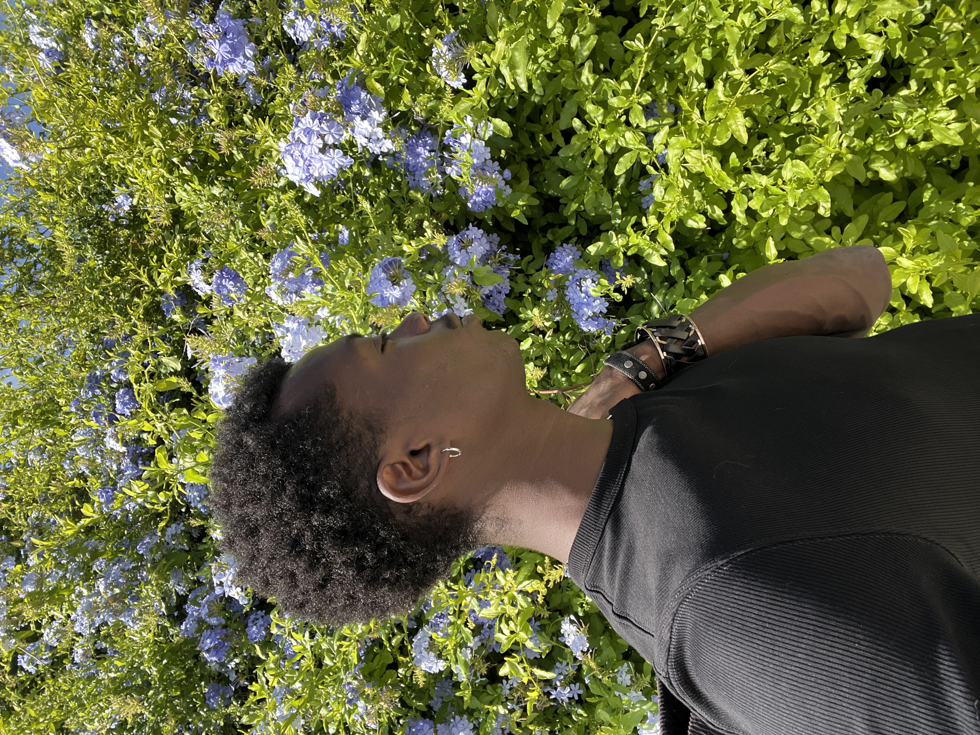 A young Black person with dark, curly hair lies on their side amongst a lush green bush with small blue flowers. The person is wearing a black t-shirt with a ribbed texture on the sleeve and chest. They have a silver hoop earring in their left ear and a brown leather bracelet with silver studs on their right wrist. Their eyes are closed, and their head is tilted back, suggesting a moment of relaxation or contemplation. The bright sunlight indicates it is daytime. The foliage and flowers suggest a natural, outdoor setting, likely a garden or park. The location context of Sant Adrià de Besòs, Spain, places this scene in a Mediterranean environment. The overall mood is peaceful and serene. There is no visible text in the image.