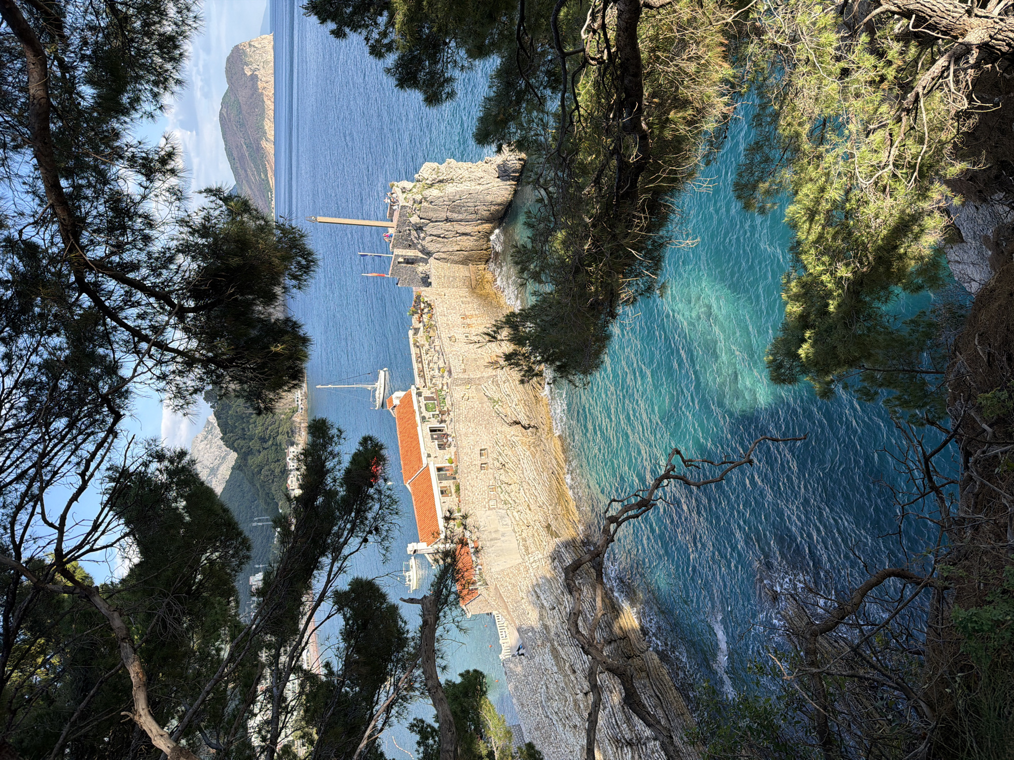 The scene is an elevated, angled view in Petrovac centar, Montenegro. In the distance is an island. The focus of the image is a rocky, fortified peninsula jutting into the turquoise sea, topped with a structure that appears to be a building with a red tile roof. A flag stands high near its tip. The peninsula is accessed by a narrow, stone walkway. There are yachts anchored. Trees frame the view. The scene is bathed in sunlight.