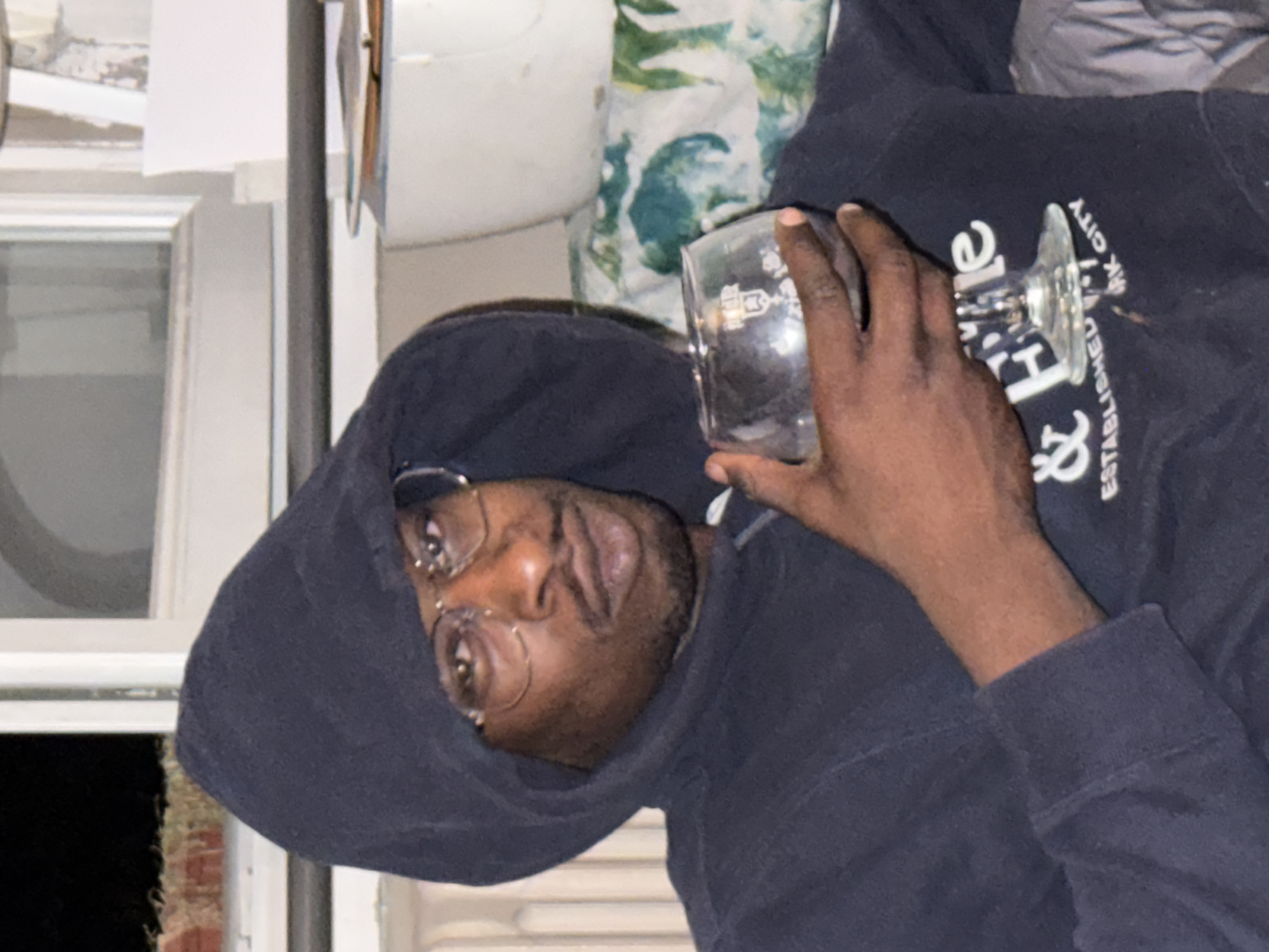 A dark-skinned person with facial hair and wire-rimmed glasses, wearing a dark hooded sweatshirt, holds a clear glass with a stem. The person is partially framed by a window with a view to the outside at night. The background includes a wall and a patterned cushion or pillow. The location information is not discernible from this image. It is not possible to verify if it is in Sarcelles, France from this image.